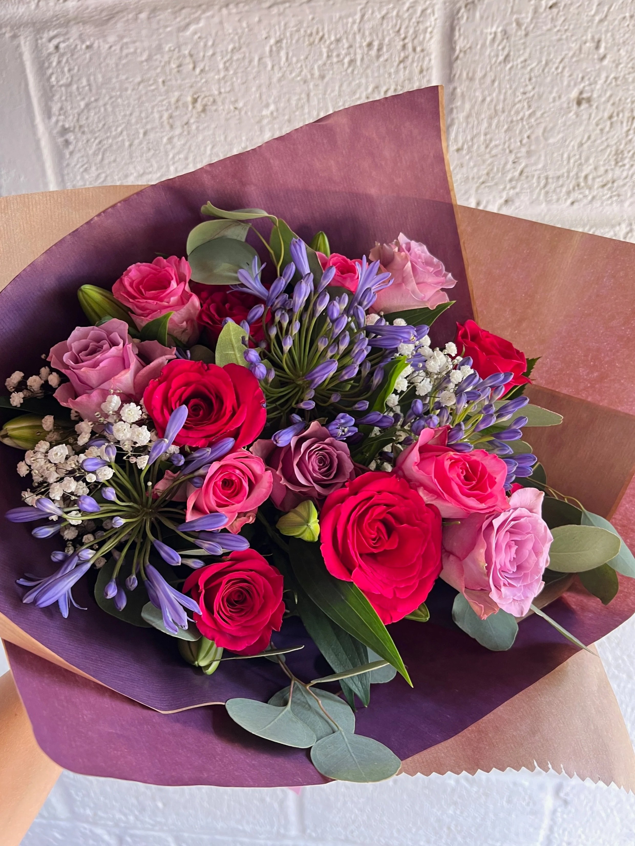 Bouquet of pink, red, and purple roses, purple agapanthus, white baby's breath, and green foliage wrapped in purple and brown paper.