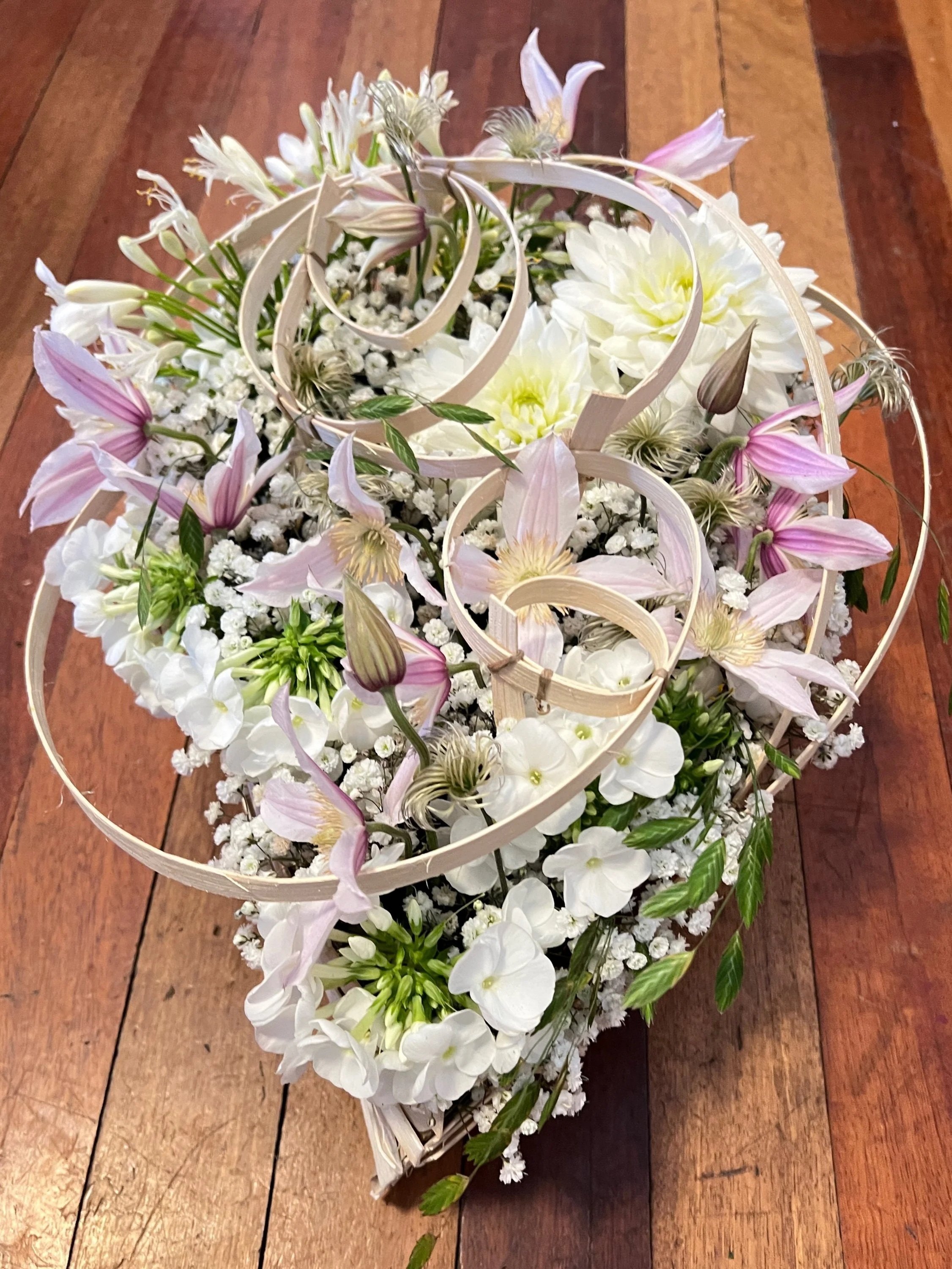 A flower arrangement with white and light pink flowers, some greenery, in a white decorative basket on a wooden surface.