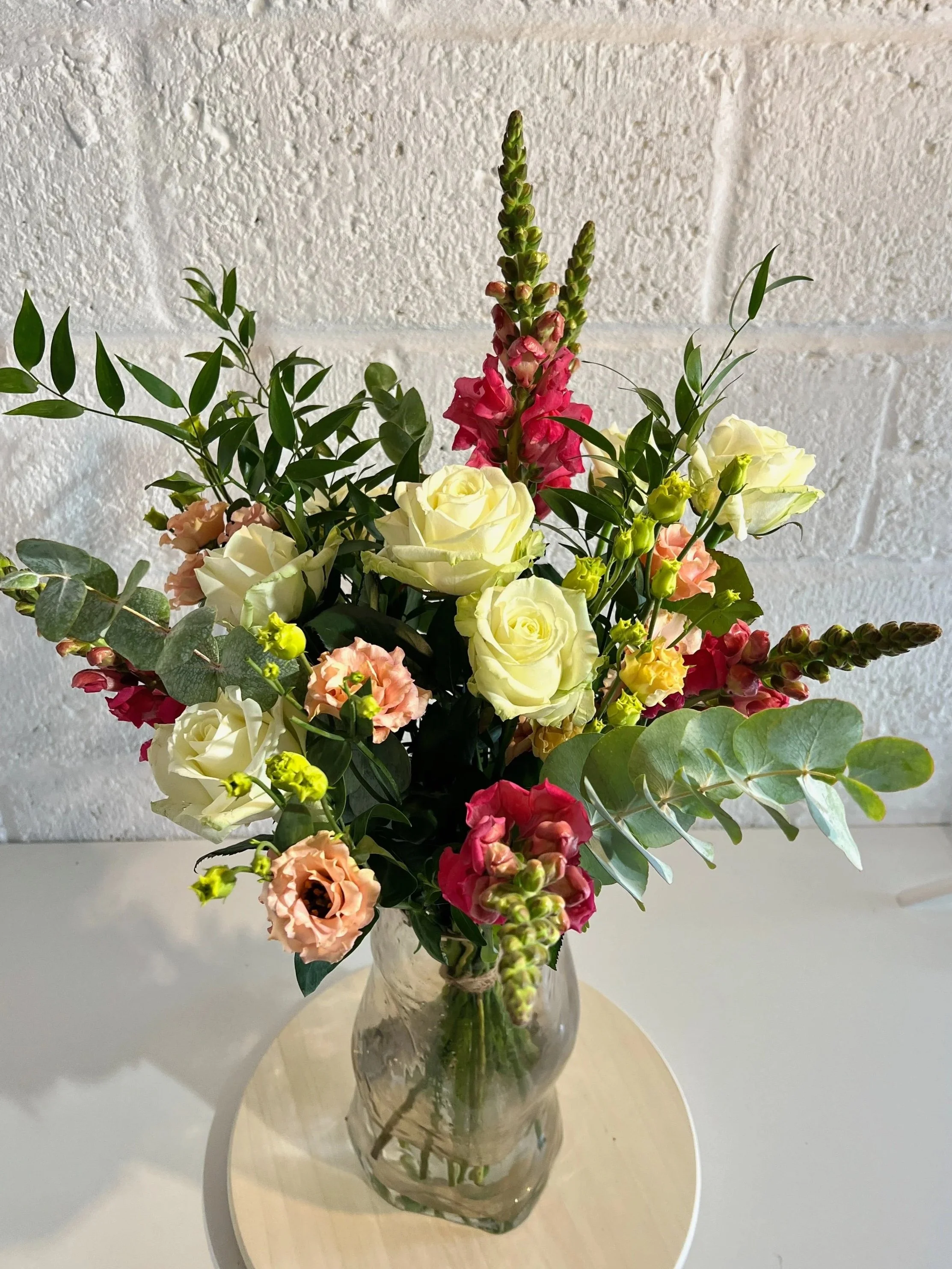 A glass vase filled with a colorful bouquet of flowers, including white roses, pink lisianthus, yellow snapdragons, and various green foliage, set on a wooden surface against a white brick wall background.