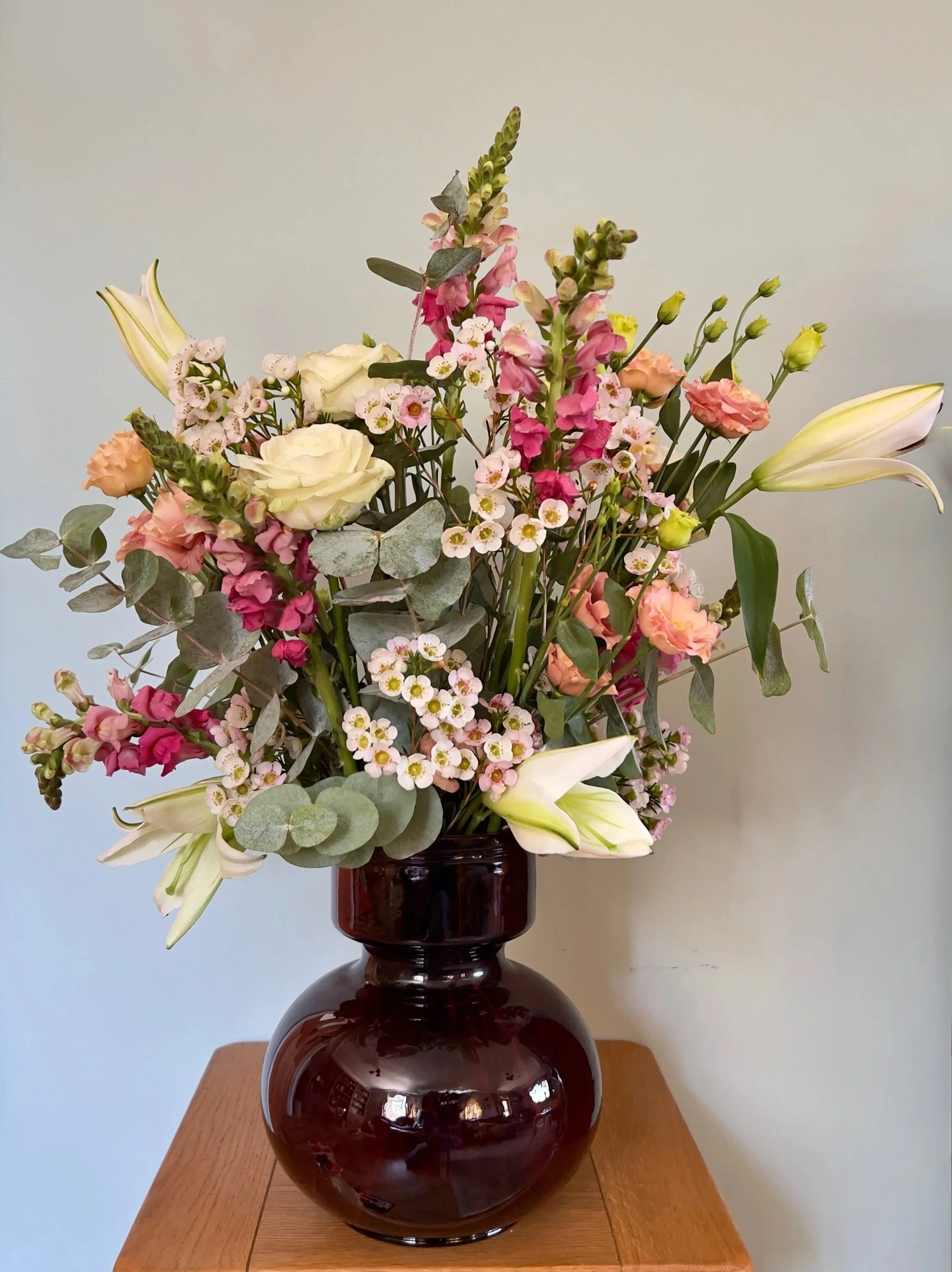 A floral arrangement in a dark round vase on a wooden table, featuring white lilies, pink and peach roses, white small flowers, and green foliage against a plain background.