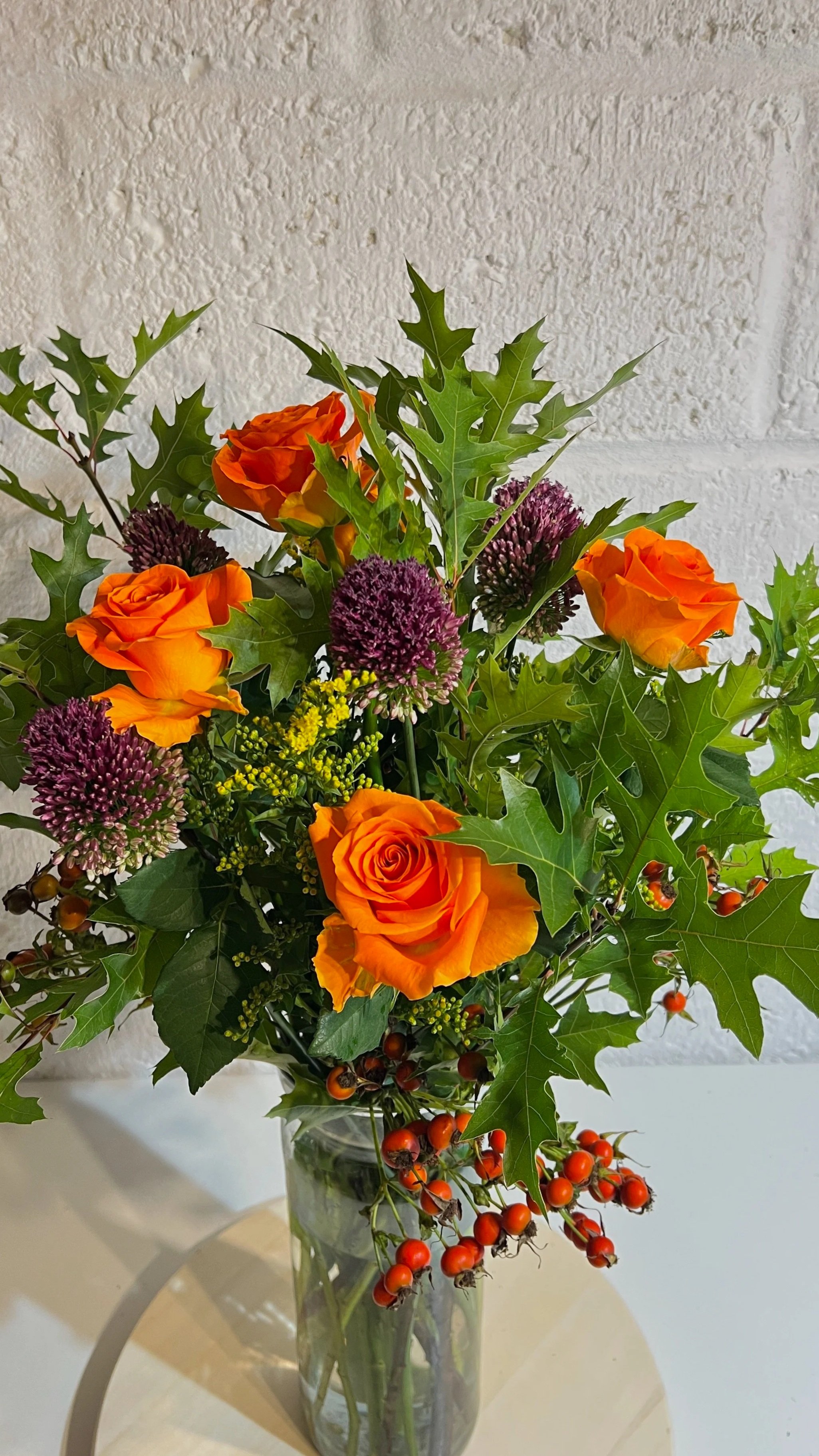 A flower arrangement with orange roses, purple allium, yellow accents, large green holly leaves, and red berry sprigs in a tall glass vase.