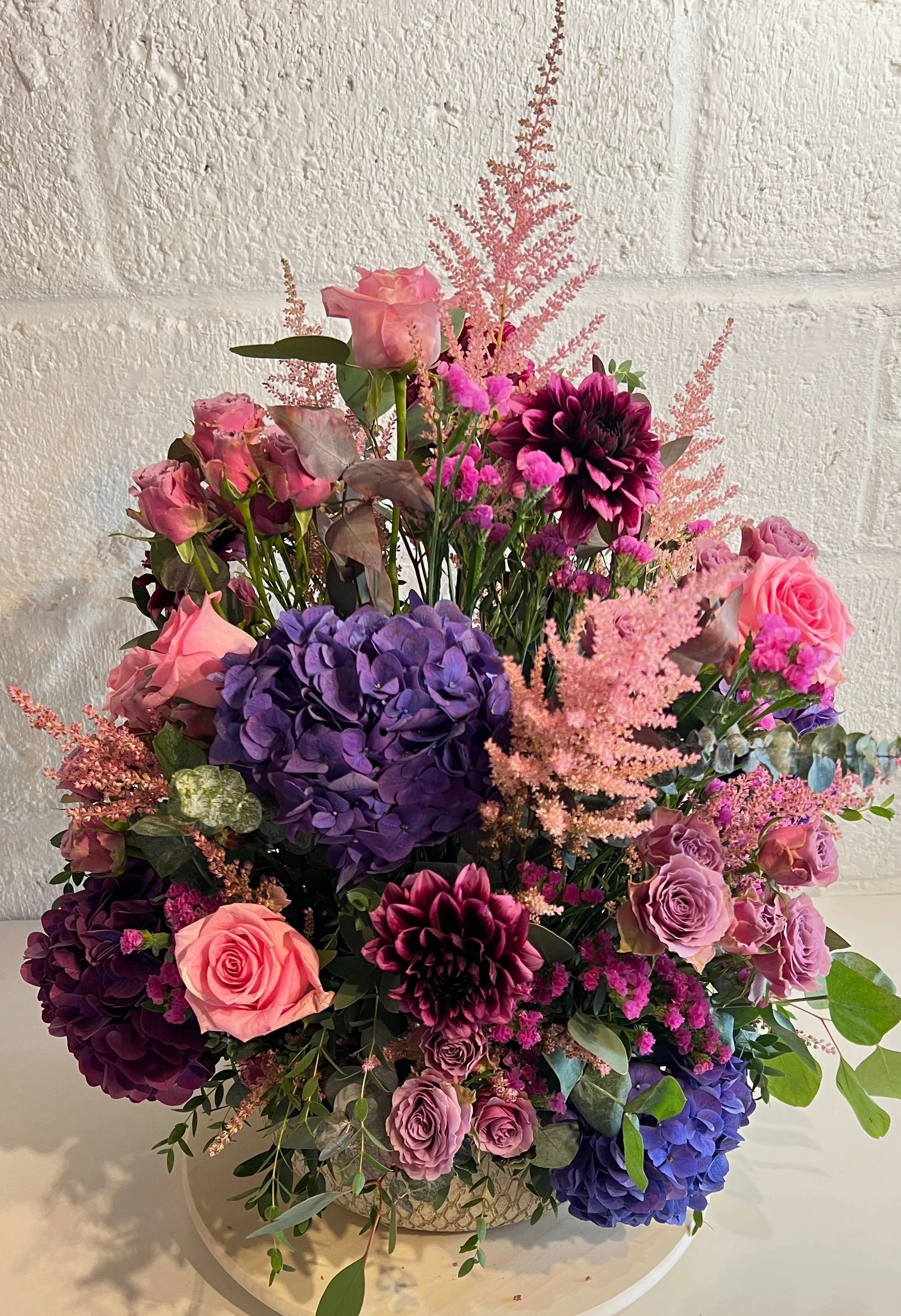 A colorful bouquet of pink, purple, and dark maroon flowers with green leaves in a round white vase.