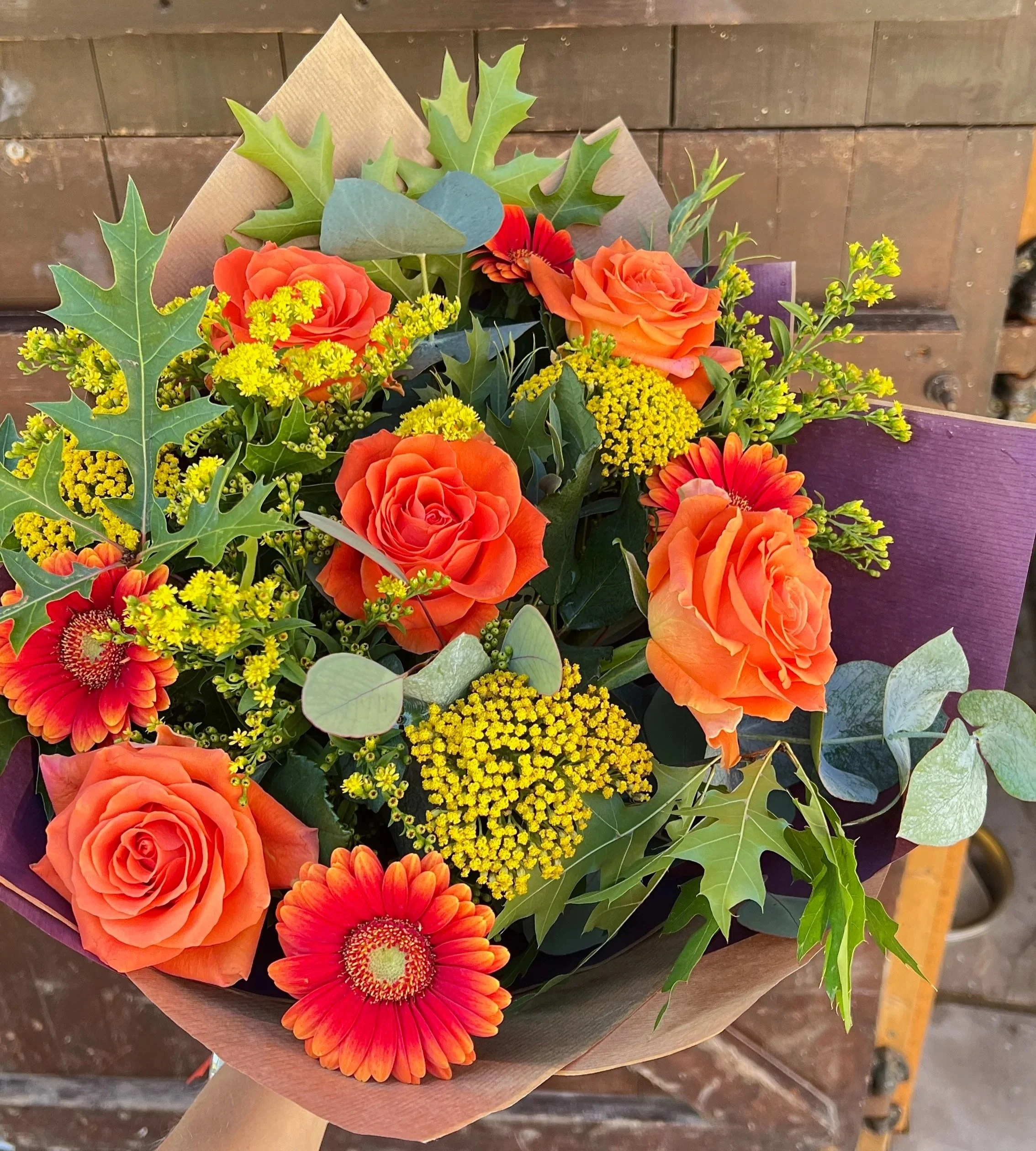 A bouquet of vibrant orange roses, red gerbera daisies, yellow filler flowers, and green foliage wrapped in brown paper, resting on a wooden surface.