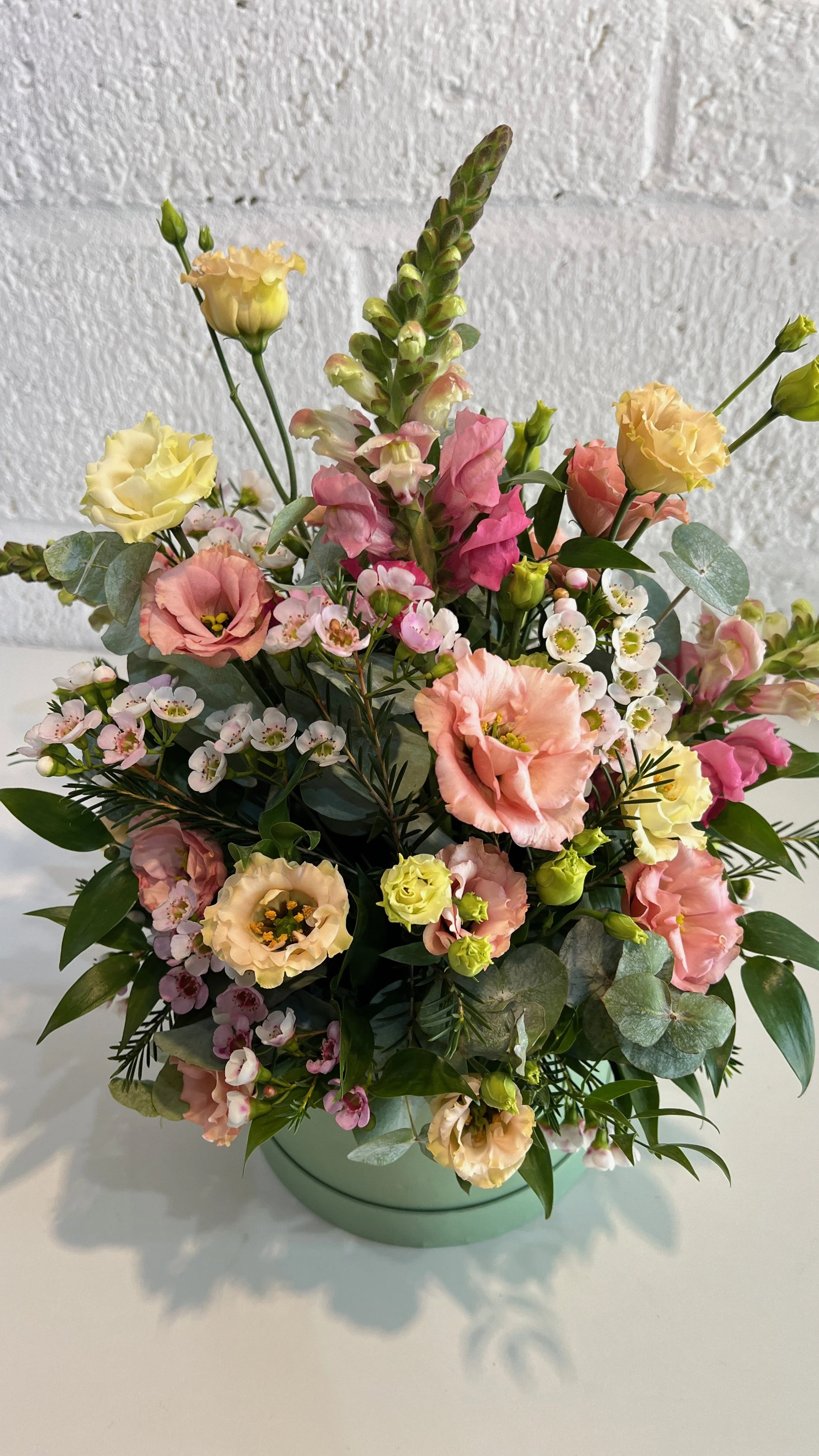 A colorful flower bouquet in a mint green vase containing pink, yellow, and cream-colored flowers along with greenery, placed on a white surface against a white textured wall.
