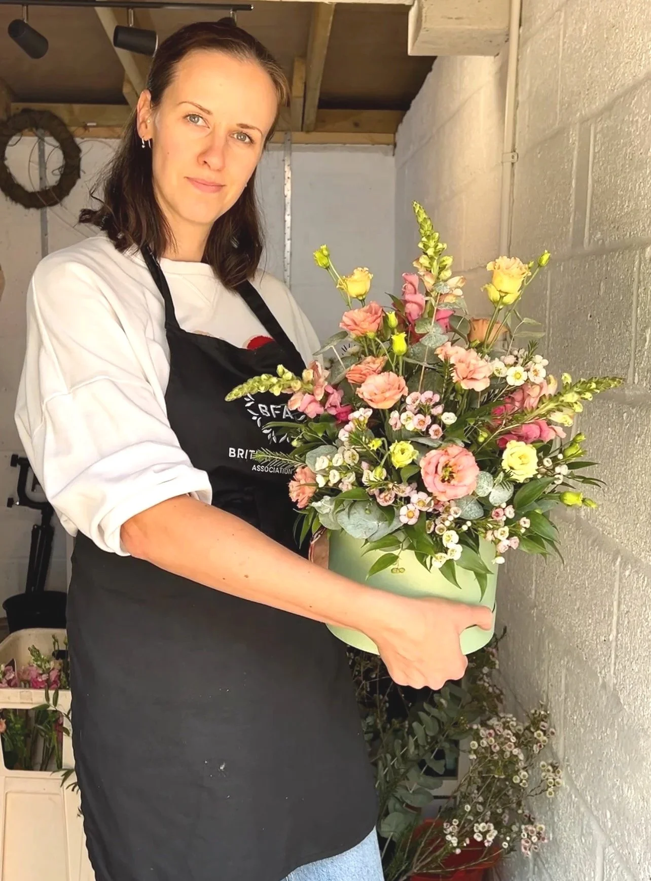 A woman holding a large, colorful floral bouquet in a green pot, standing in a workshop or flower shop.