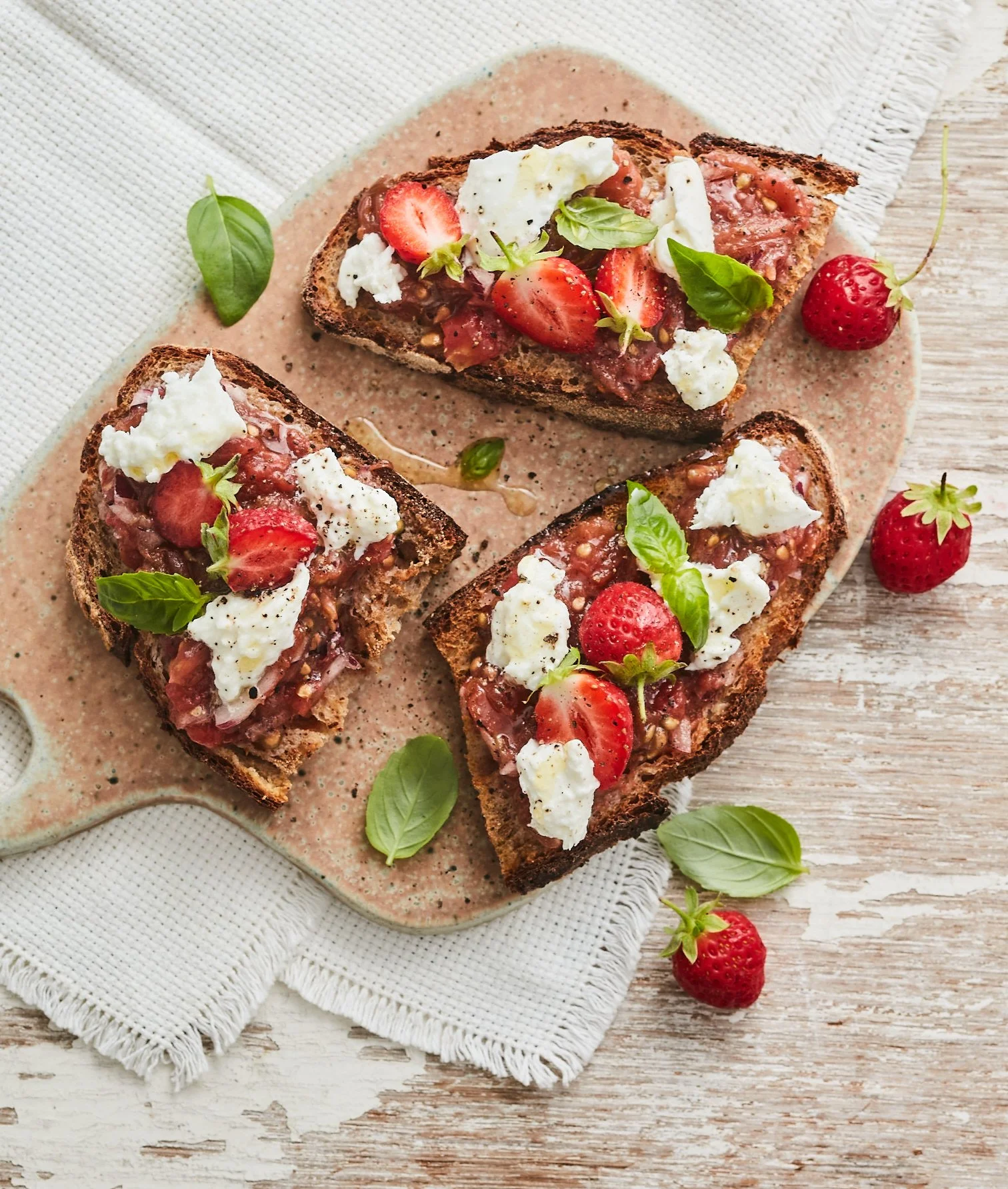 Tartines de pain grillé garnies de frais, de fraises, de basilic et de fromage, sur une planche en bois avec quelques fraises et feuilles de basilic autour.