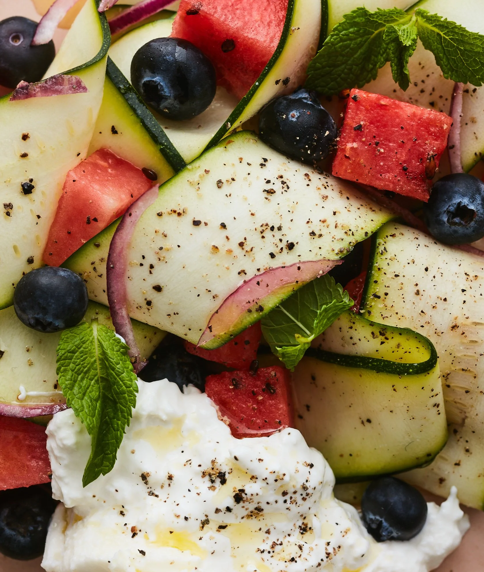 Un plat de fruits frais avec morceaux de pastèque, melon, tomate, myrtilles, et feuilles de menthe, garni de poivre noir.