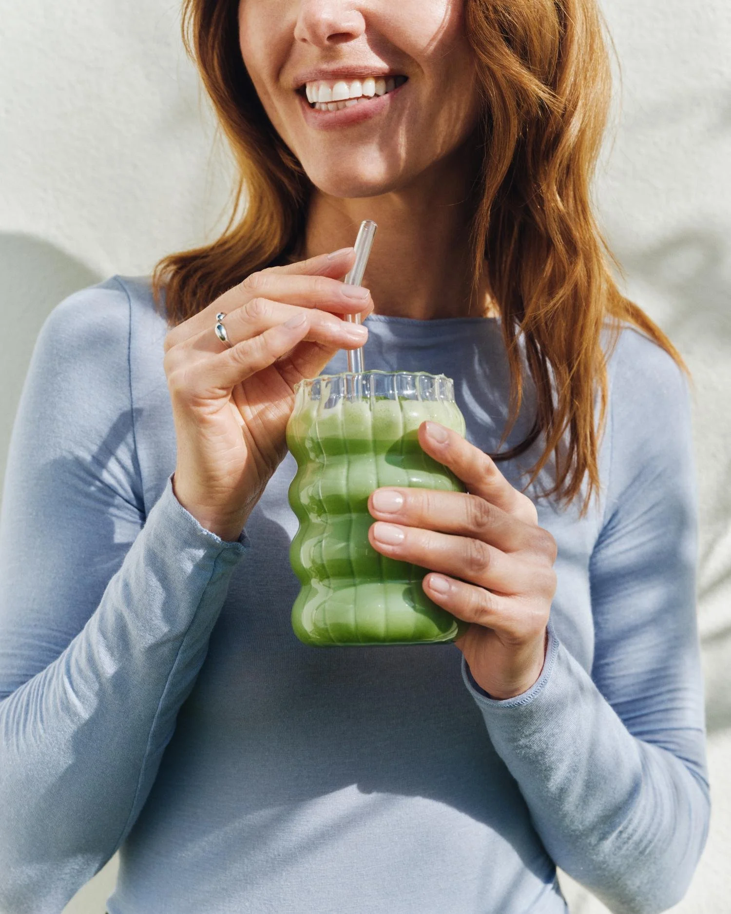 Femme avec cheveux roux buvant une boisson verte dans un verre ondulé.