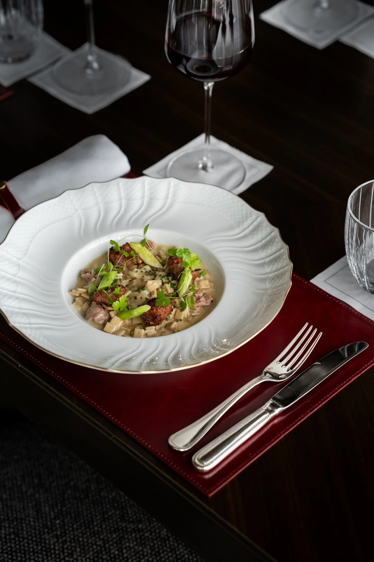 Une assiette de repas avec des pâtes, garnie de morceaux de viande, de légumes verts et de coriandre, accompagnée d'un verre de vin rouge et de vaisselle élégante sur une nappe blanche.