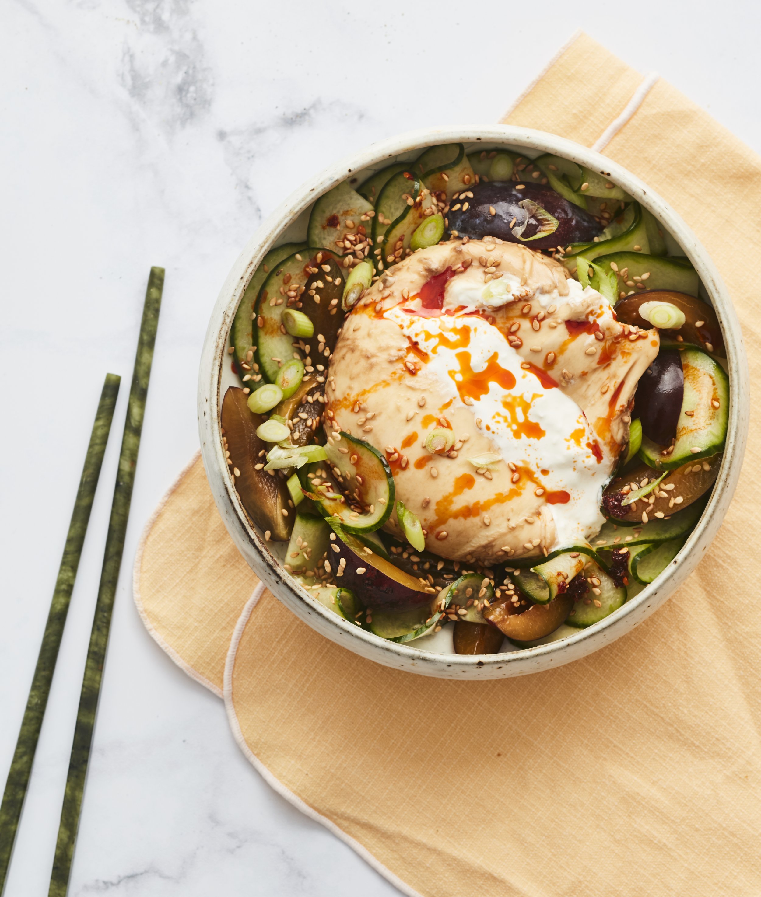 Un bol de salade avec des tranches de concombre, des œufs pochés, des graines de sésame et des morceaux d’aubergine, sur une nappe beige, avec des baguettes de cuisine à côté.