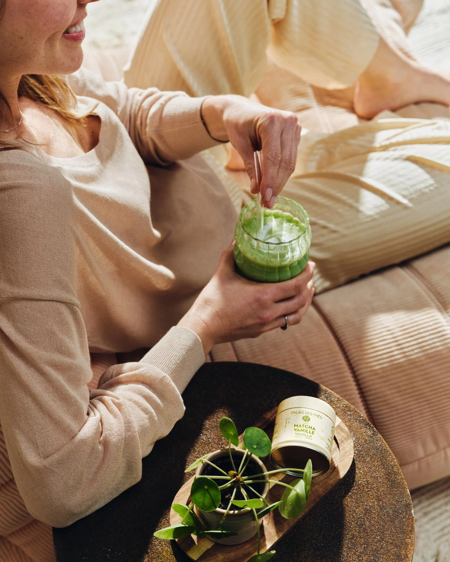 Une femme prépare une boisson dans un verre vert, avec une plante verte en pot et un pot de thé matcha vanille sur une table en bois. Elle sourit et porte un sweatshirt beige.