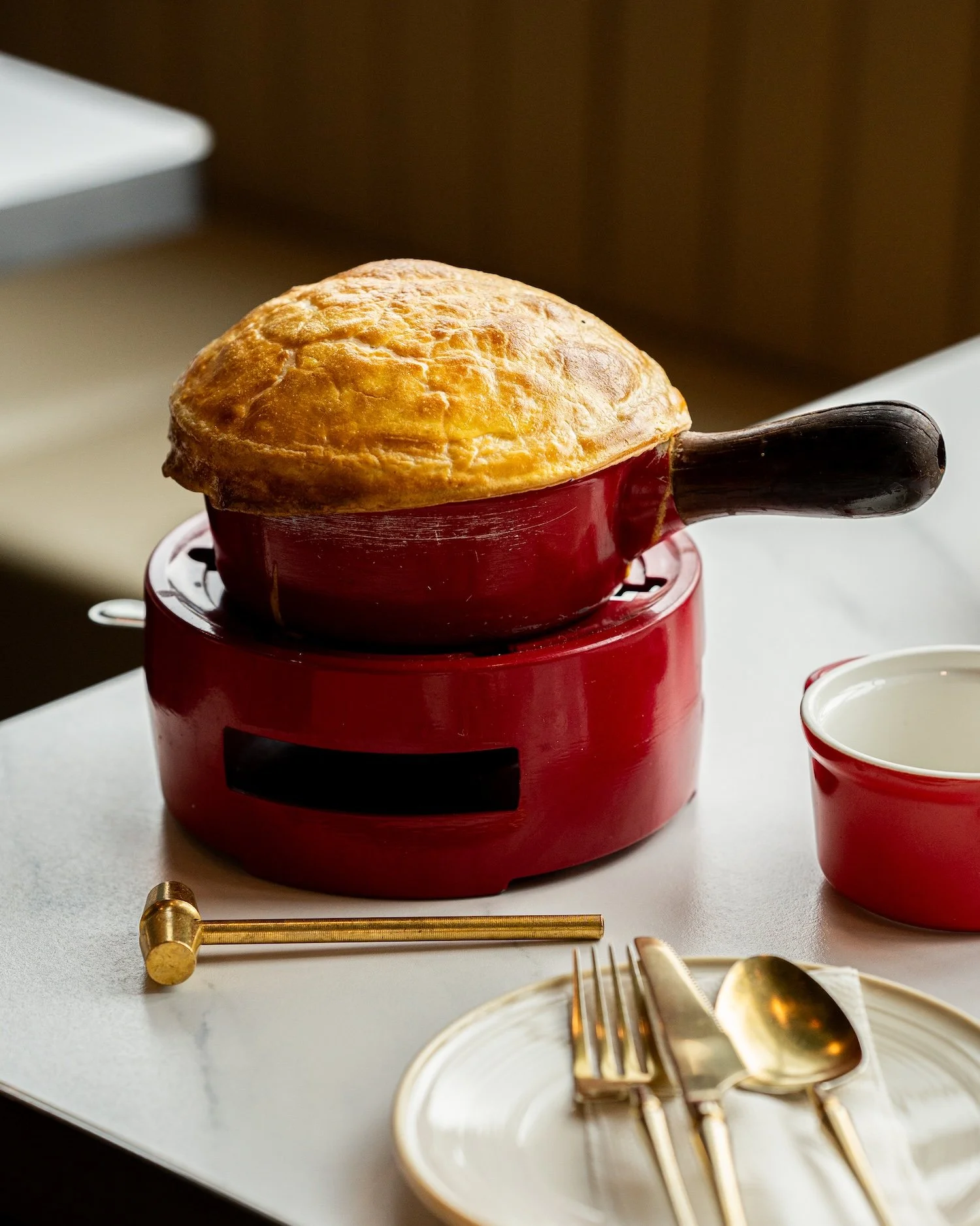 Une tarte ou un gâteau dans un bol en porcelaine rouge, posé sur un mini réchaud, avec une assiette, une fourchette, une cuillère et une baguette en or sur une table blanche, dans un décor intérieur.