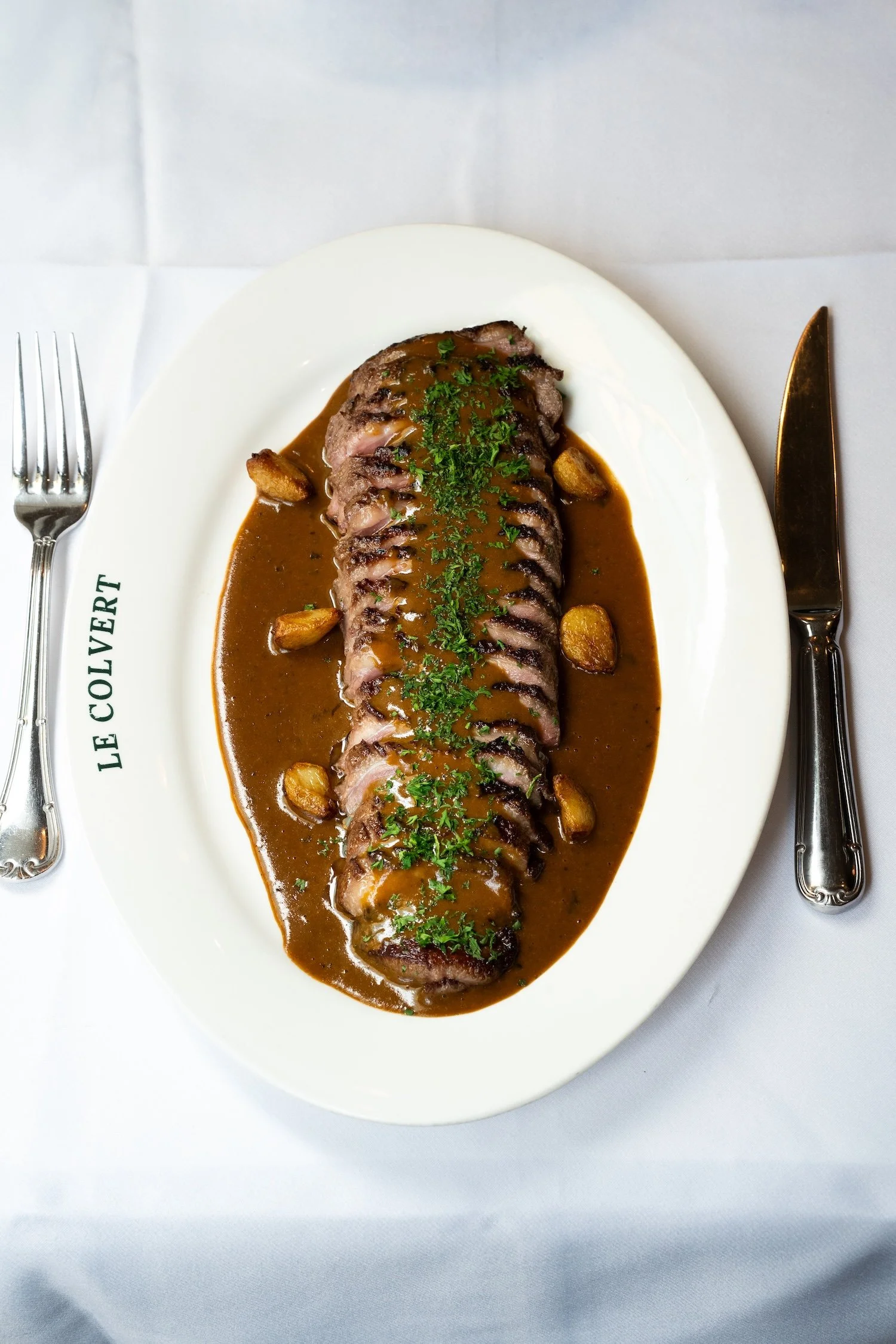 Une assiette avec un filet de bœuf présenté avec une sauce brune, garnie de persil et d'ail confit, servant de plat principal dans un restaurant.