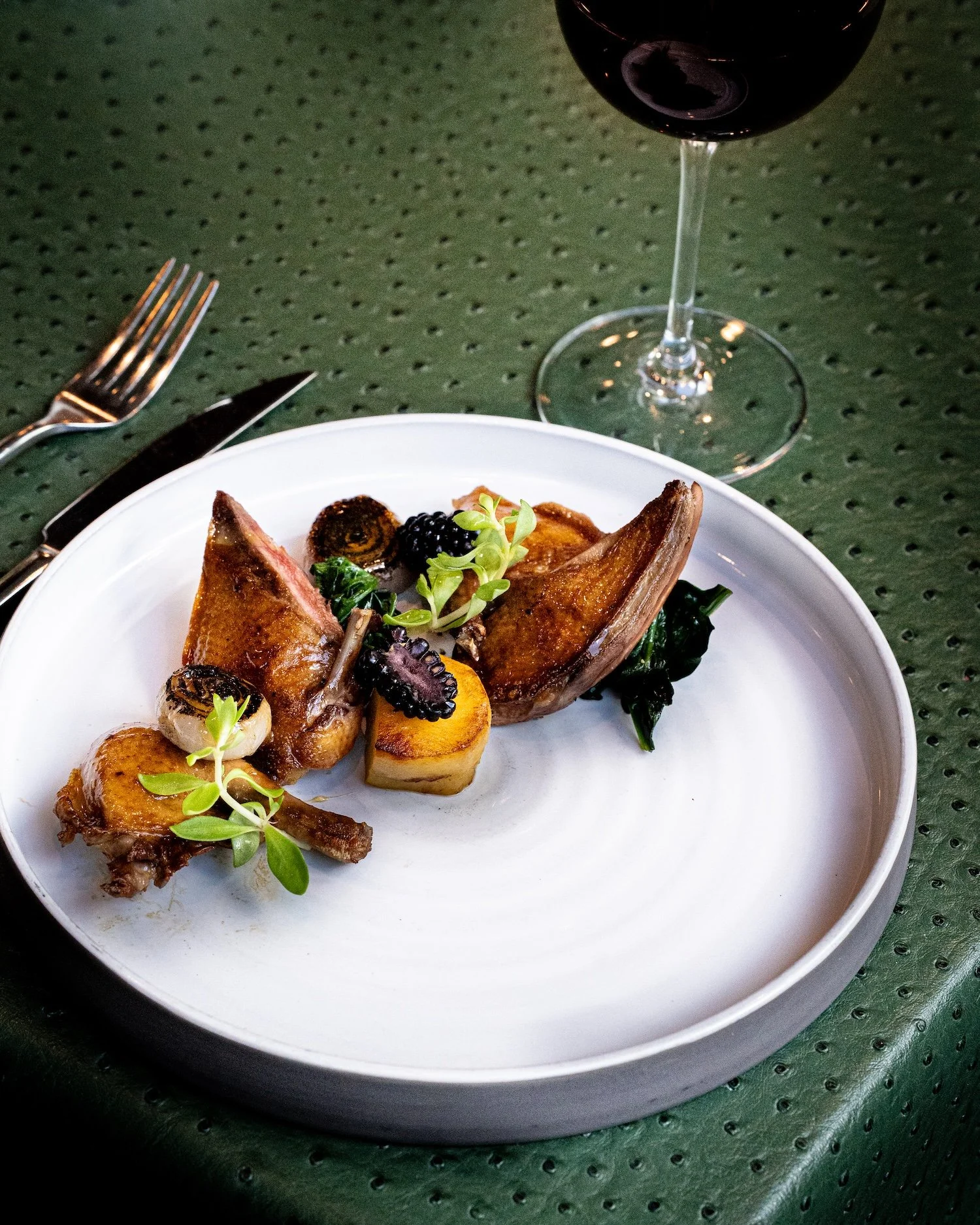 Assiette avec deux morceaux de viande, légumes, fruits noirs et garnitures, servie avec un verre de vin rouge.
