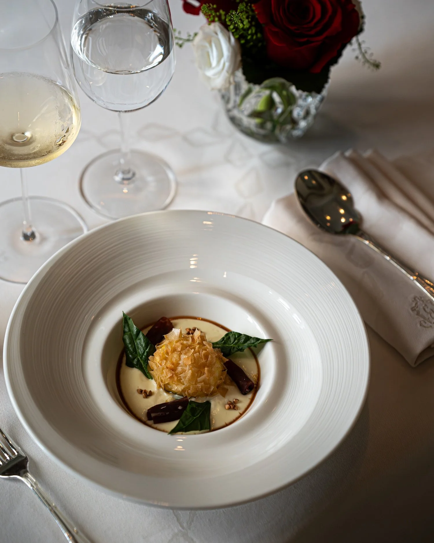 Assiette de soupe avec des garnitures d'herbes, de croûtons, et des morceaux de légumes, servie dans une assiette blanche, avec une table élégante, un bouquet de fleurs et des couverts à côté.