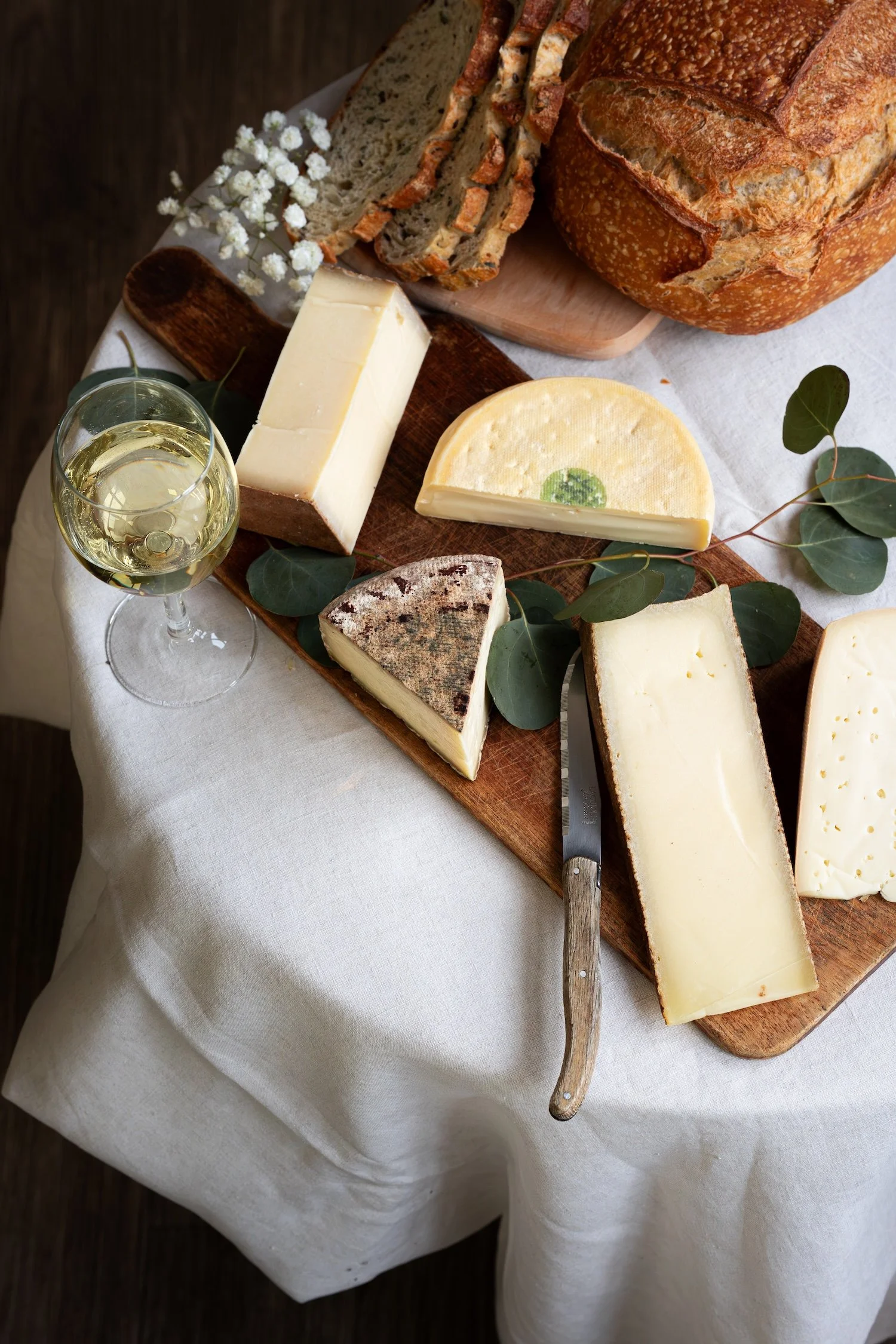 Plateau avec diverses variétés de fromages, un verre de vin blanc, une tranche de pain et des feuilles de eucalyptus, sur une table avec une nappe blanche.