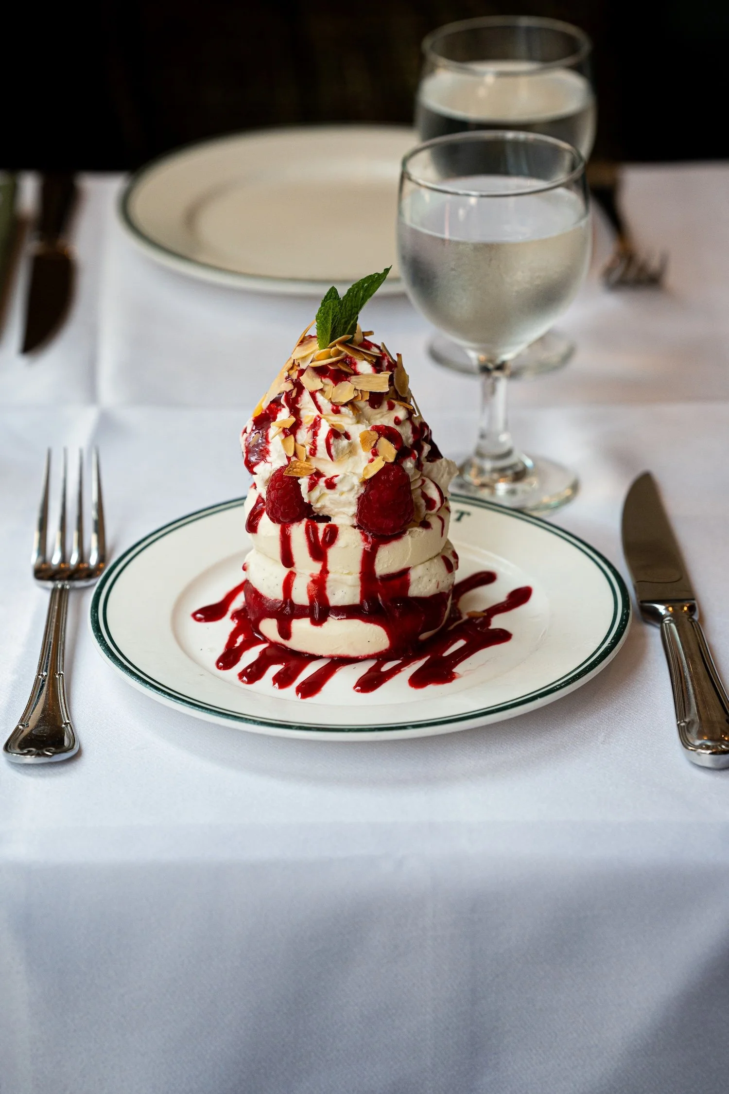 Une assiette avec un dessert, probablement une pavlova, garnie de framboises, d'amandes effilées, d'une feuille de menthe, et de coulis de framboise, accompagnée de deux verres d'eau.