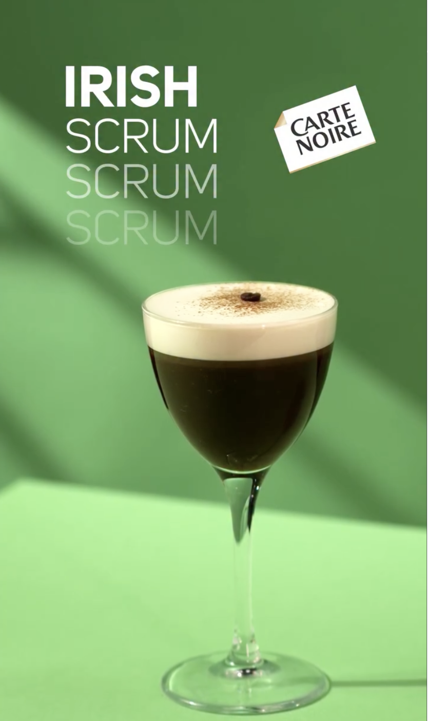Un cocktail noir dans un verre à vin avec une couche de crème au dessus, décoré d'un grain de café, sur un fond vert avec du texte mentionnant 'Irish Scrum Scrum' et une étiquette 'Carte Noire'.