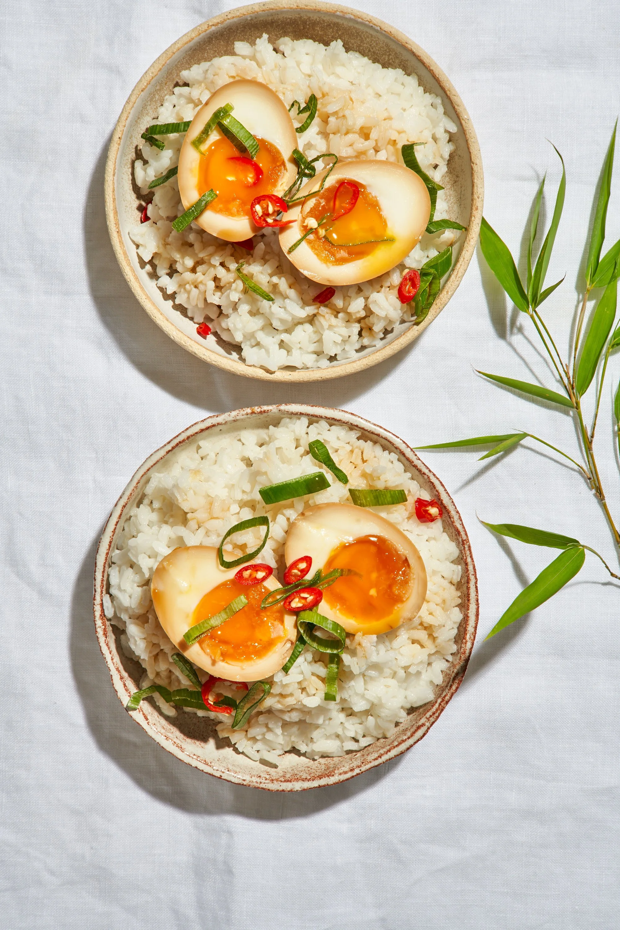 Bol de riz avec œuf mollet coupé en deux, garni de tranches de piment rouge et de ciboulette émincée, sur une nappe blanche avec quelques brins de bambou à côté.