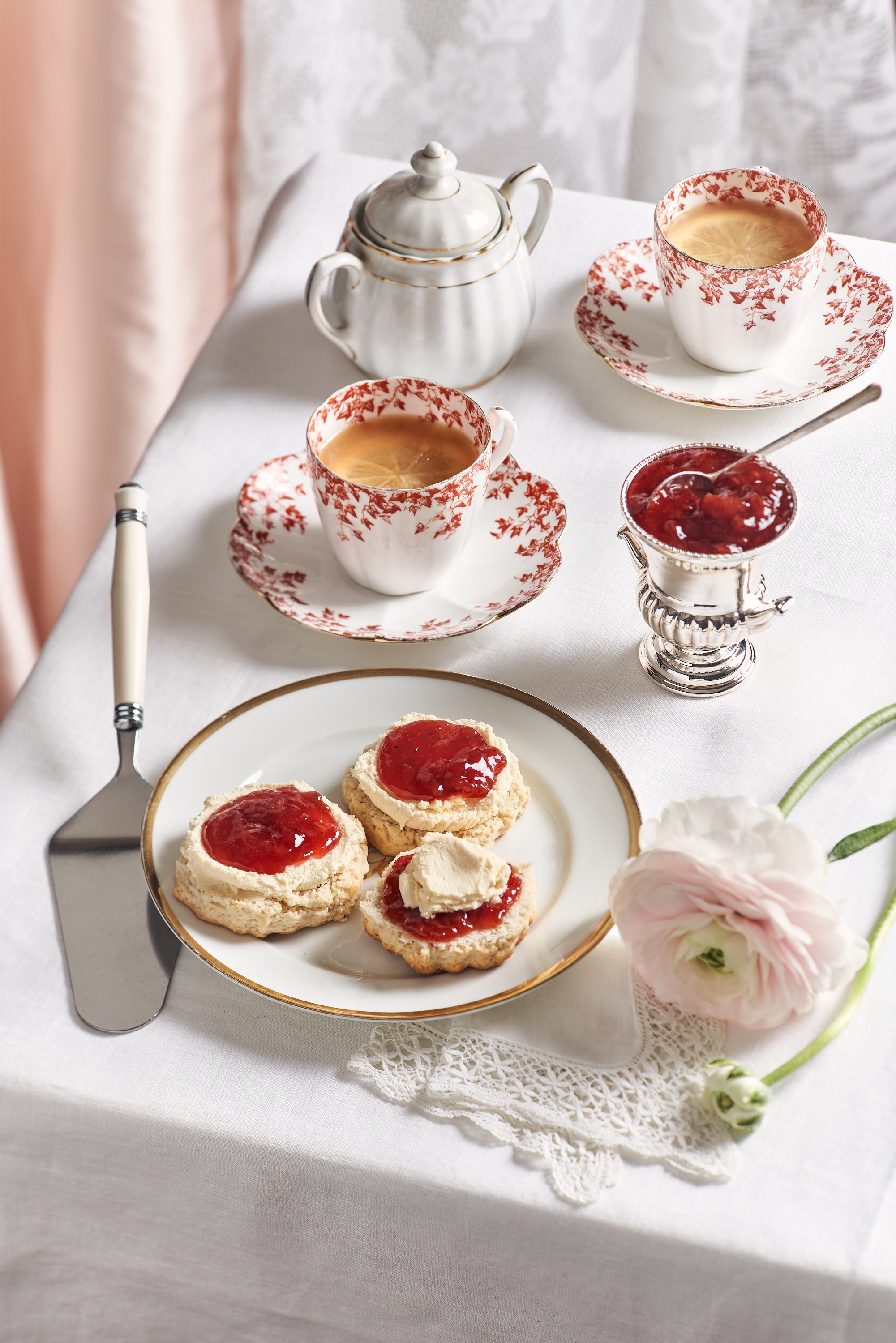 Une tasse de thé ou de café avec de la crème ou du lait, une théière blanche, une petite coupe à sauce avec de la confiture, une assiette avec des biscuits tartinés de confiture, une fleur de pivoine pâle sur une dentelle blanche, et un couteau avec une poignée blanche et métallique. Le tout est posé sur une nappe blanche