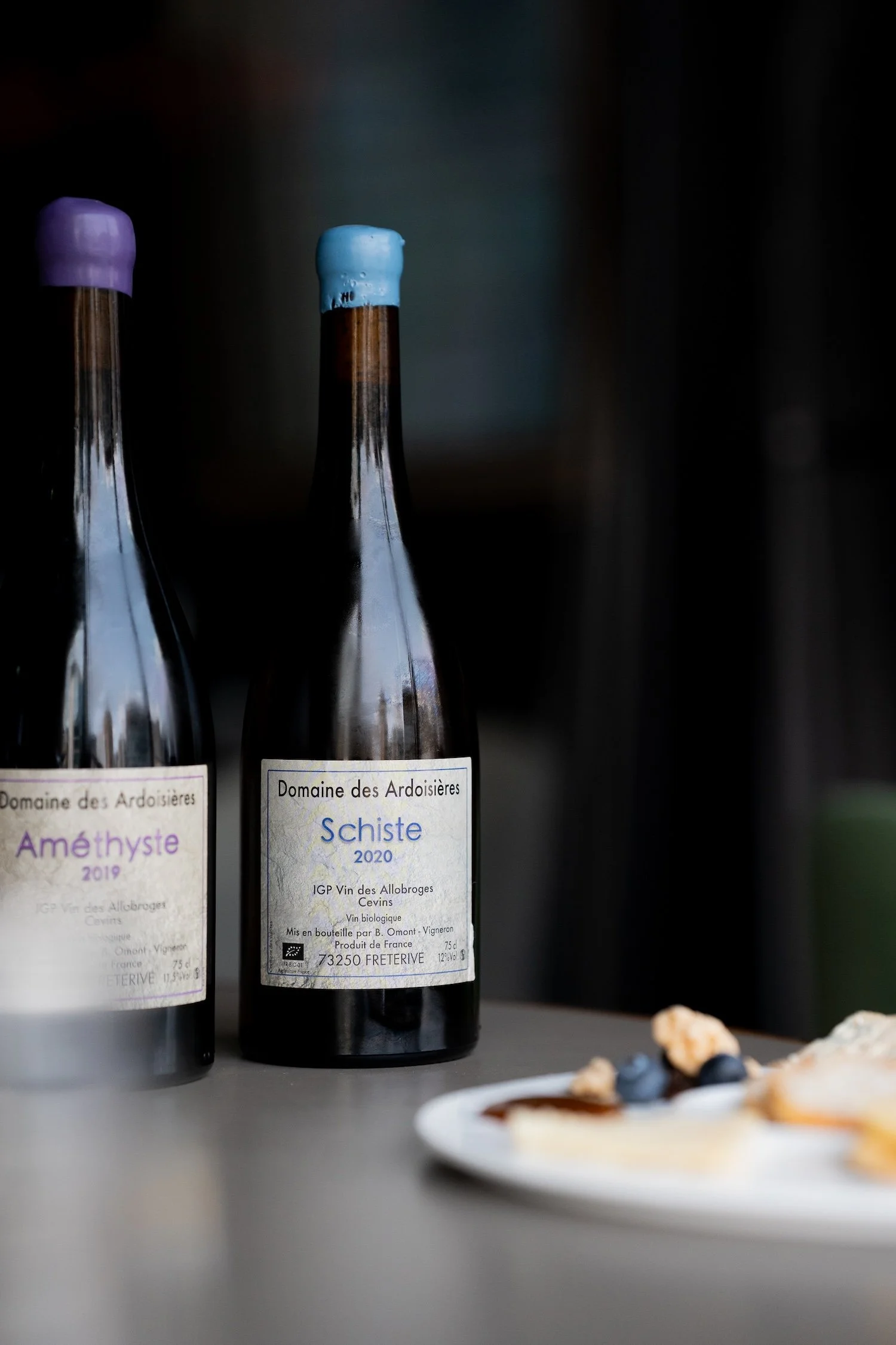 Deux bouteilles de vin rouge avec des étiquettes en français, une de 2019 et l'autre de 2020, sur une surface grise. À droite, une assiette avec un dessert blanc, confiture ou sauce, et des myrtilles.