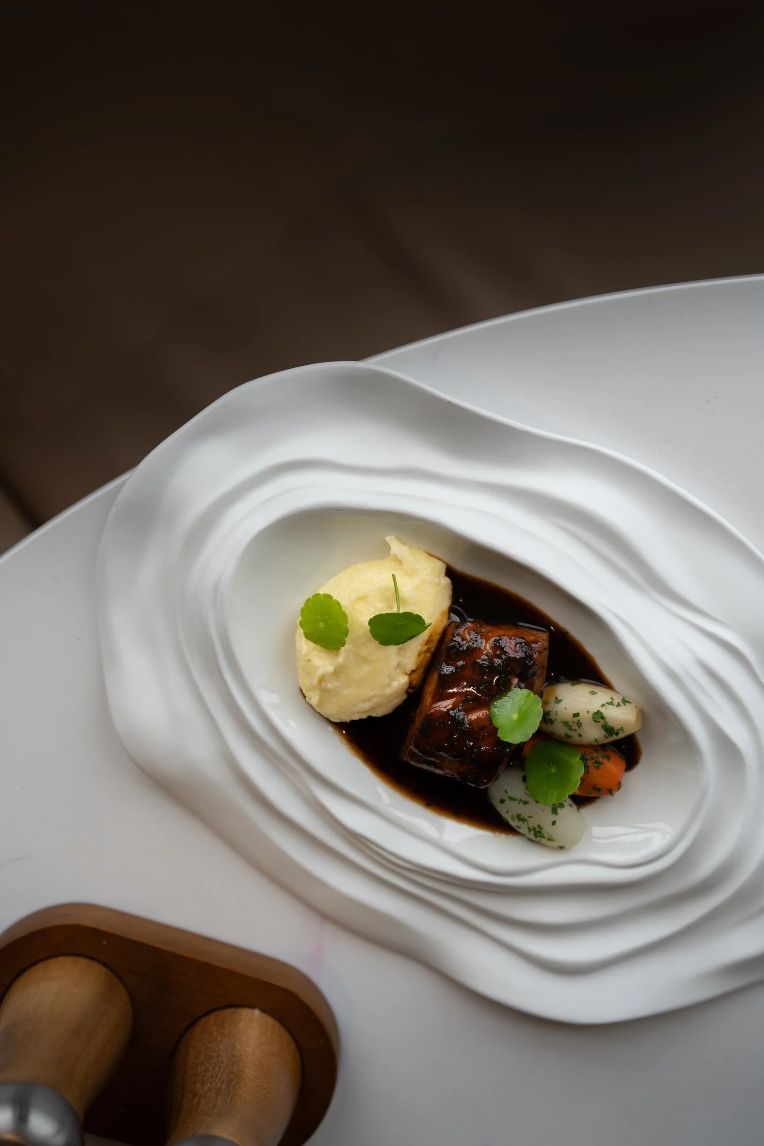 Présentation d'un plat gastronomique dans une assiette blanche avec un filet de poisson, une purée, des légumes, et une sauce foncée, décoré de feuilles de cilantro.