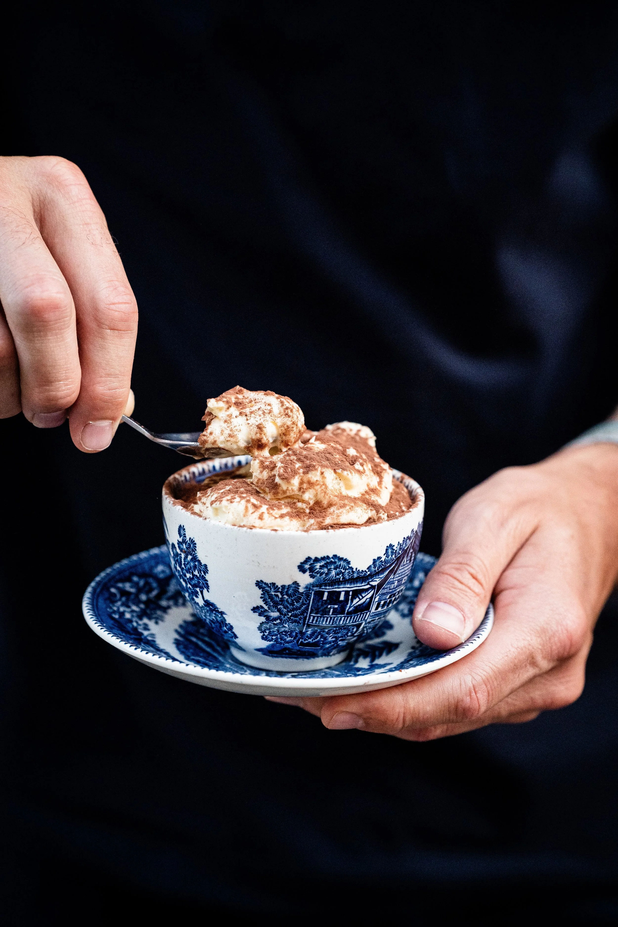 Une personne tient une tasse avec un dessert crémeux saupoudré de cacao. La tasse est en porcelaine avec un motif bleu et blanc, placée sur une petite soucoupe assortie, le fond est noir.