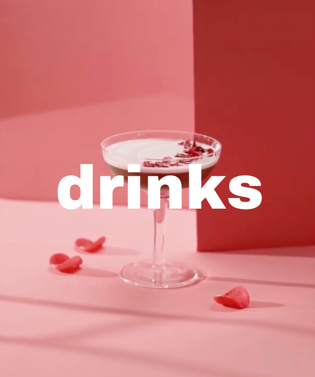 Un verre à cocktail avec une boisson blanche et des morceaux de garniture en haut, posé sur une surface rose avec quelques bonbons roses autour, sur un fond avec des nuances de rose. Le mot 'drinks' est écrit en blanc au centre de l'image.