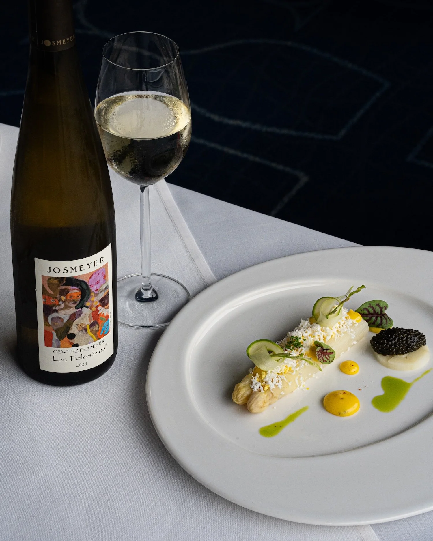Bouteille de vin blanc, verre de vin blanc, assiette avec un plat gastronomique comprenant des asperges, des sauces et des garnitures, napperon blanc, fond sombre du sol