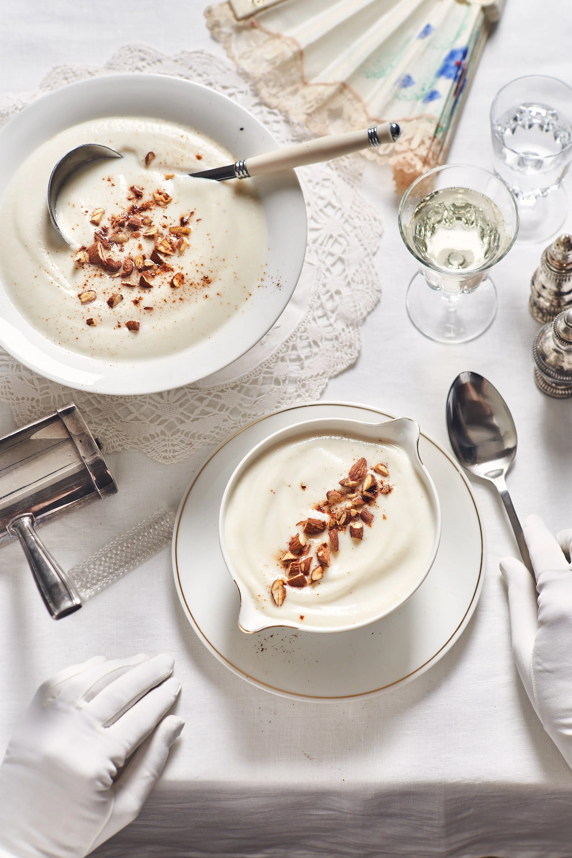 Crème dessert dans un bol blanc garni de chocolat râpé, avec une cuillère, un verre de vin blanc, un verre d'eau, des épices, et un livre ancien en arrière-plan sur une nappe en dentelle.