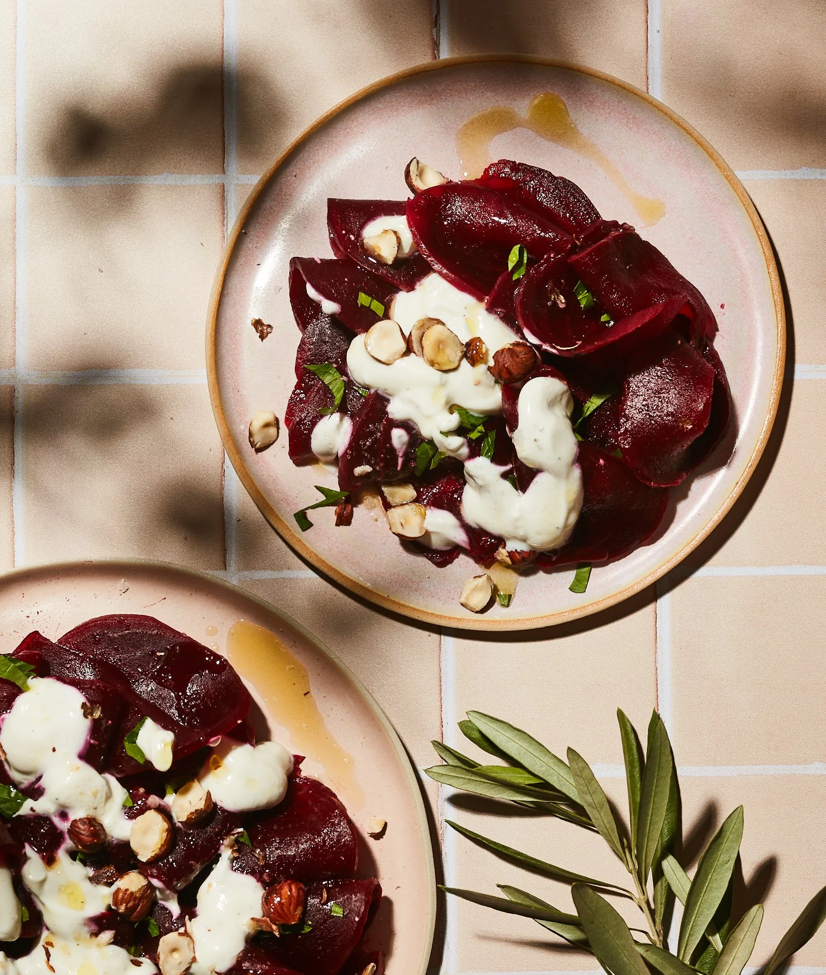 Une assiette contenant des tranches de betteraves rouges, de la crème, des noisettes, et des herbes vertes, avec une branche de feuilles vertes à côté.