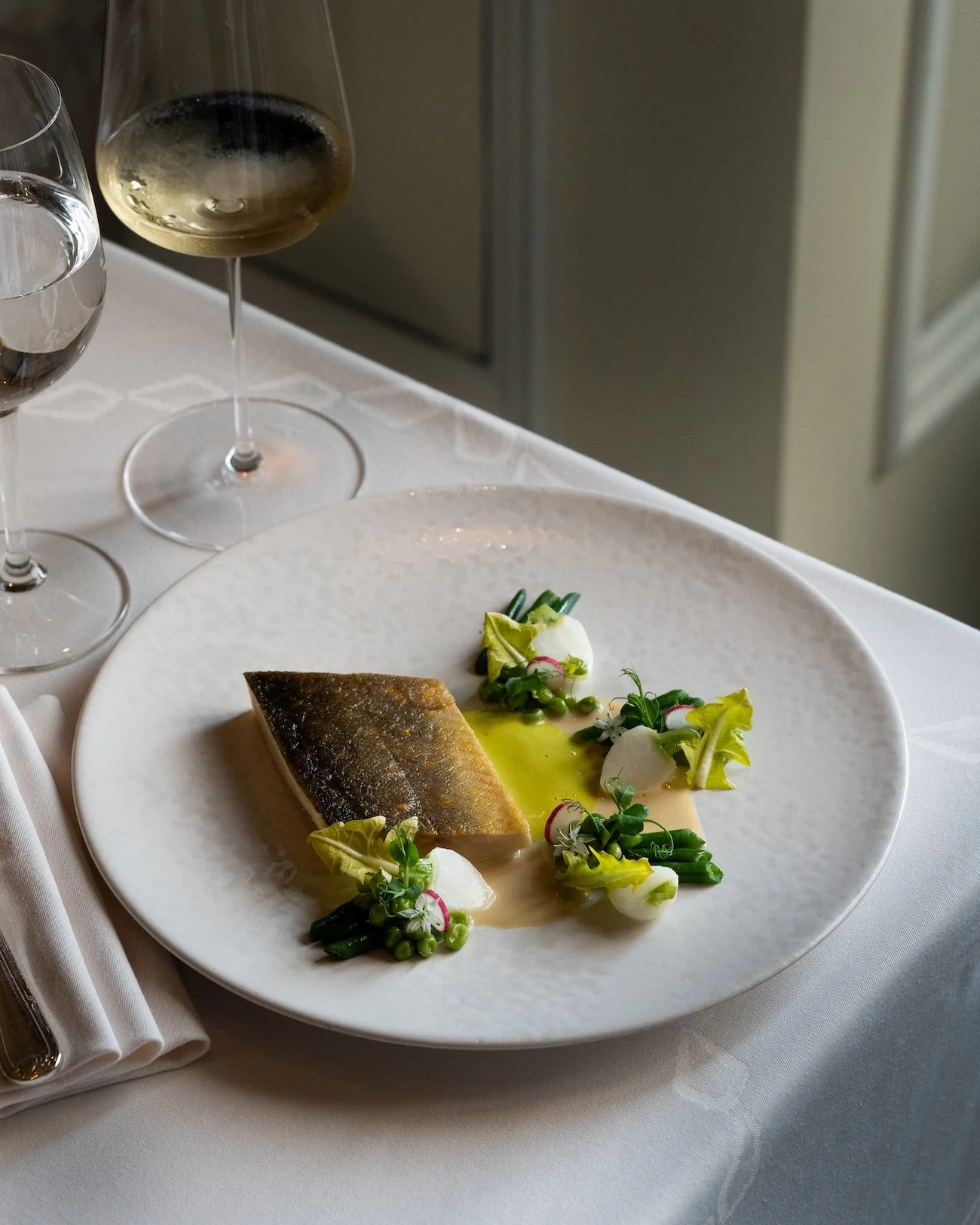 Un plat de poisson avec des légumes verts, des radis et une sauce verte, accompagné de deux verres de vin blanc sur une nappe blanche.