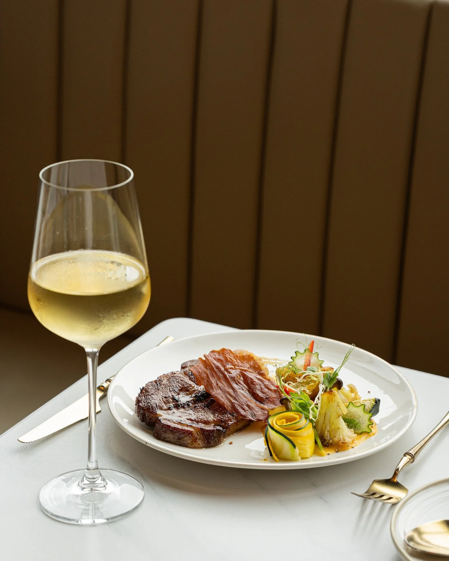Un verre de vin blanc, un plat avec une tranche de viande rouge, un morceau de poitrine de porc, et une salade ou garniture avec des légumes, sur une table élégante