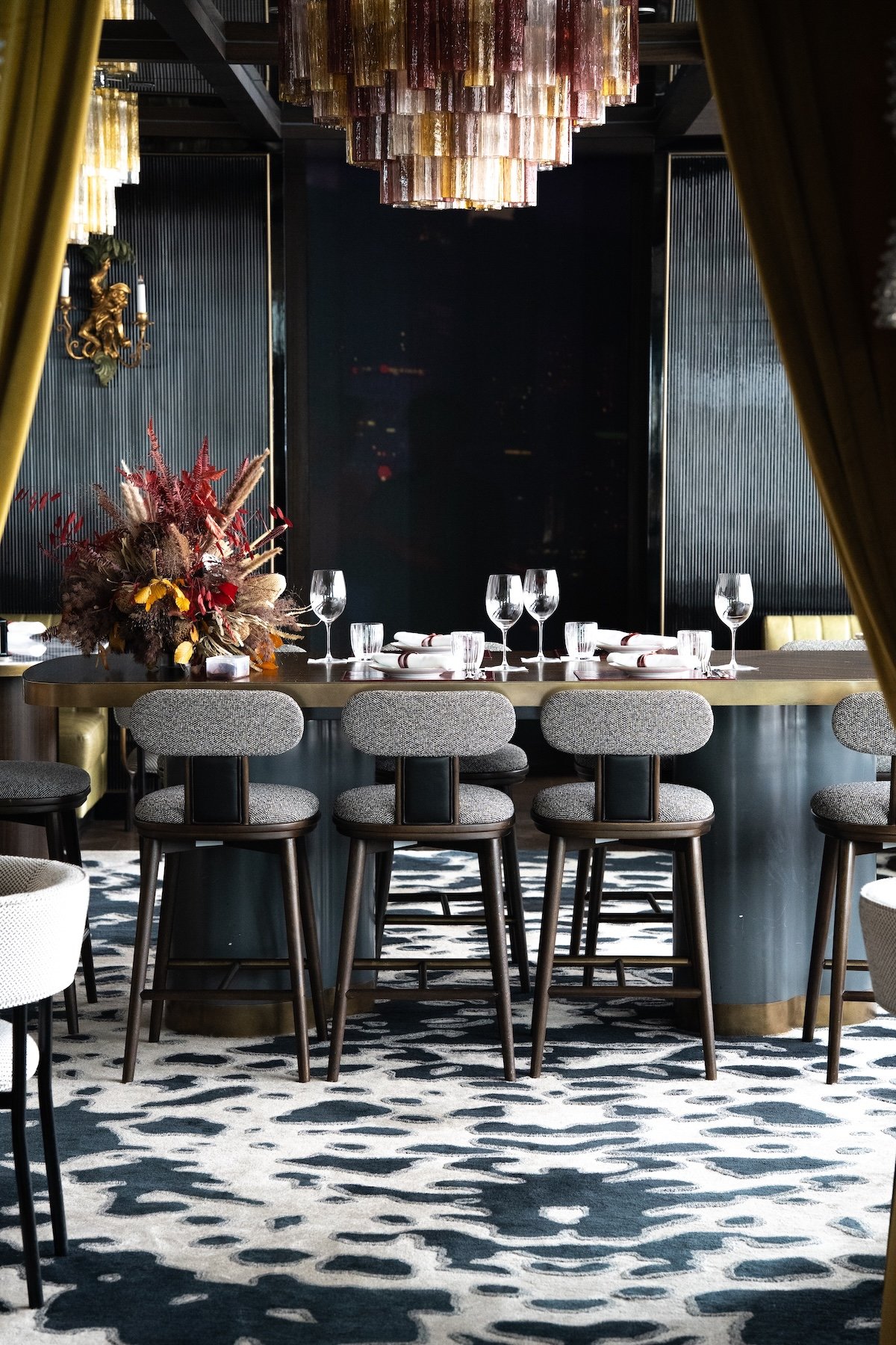 Une table élégante dans un restaurant avec des verres à vin, des assiettes, des couverts, et un gros bouquet de fleurs séchées. En arrière-plan, un mur noir avec un éclairage doré, un chandelier en verre suspendu au plafond, et des rideaux dorés.