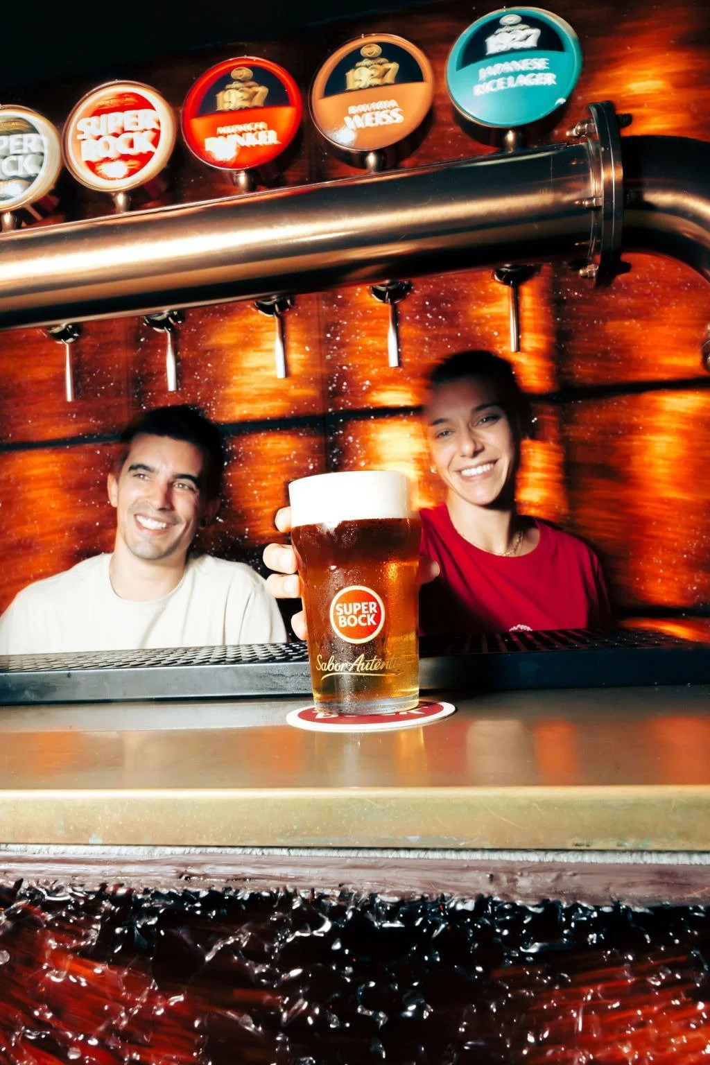 Deux personnes souriantes derrière un bar, tenant une bière Super Bock, avec des tireuses à bière en arrière-plan.