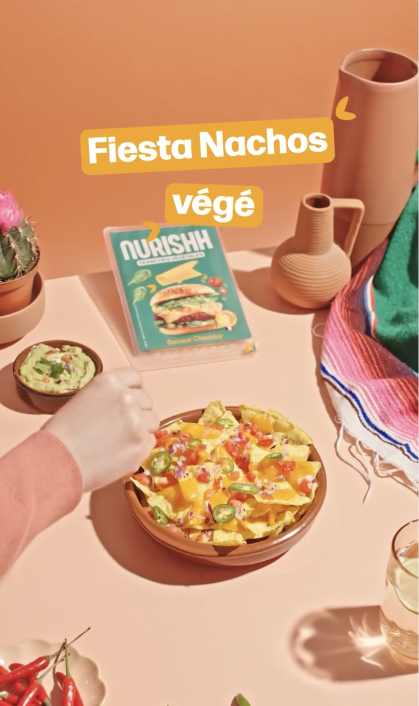 Assiette de nachos végétaliens garnis de sauce, jalapeños, tomates et autres légumes, avec un bol de guacamole à côté, sur une table rose avec décor de plantes cactées et vases en terre cuite. Marques de nachos végétaliens sur l'affiche en arrière-plan.