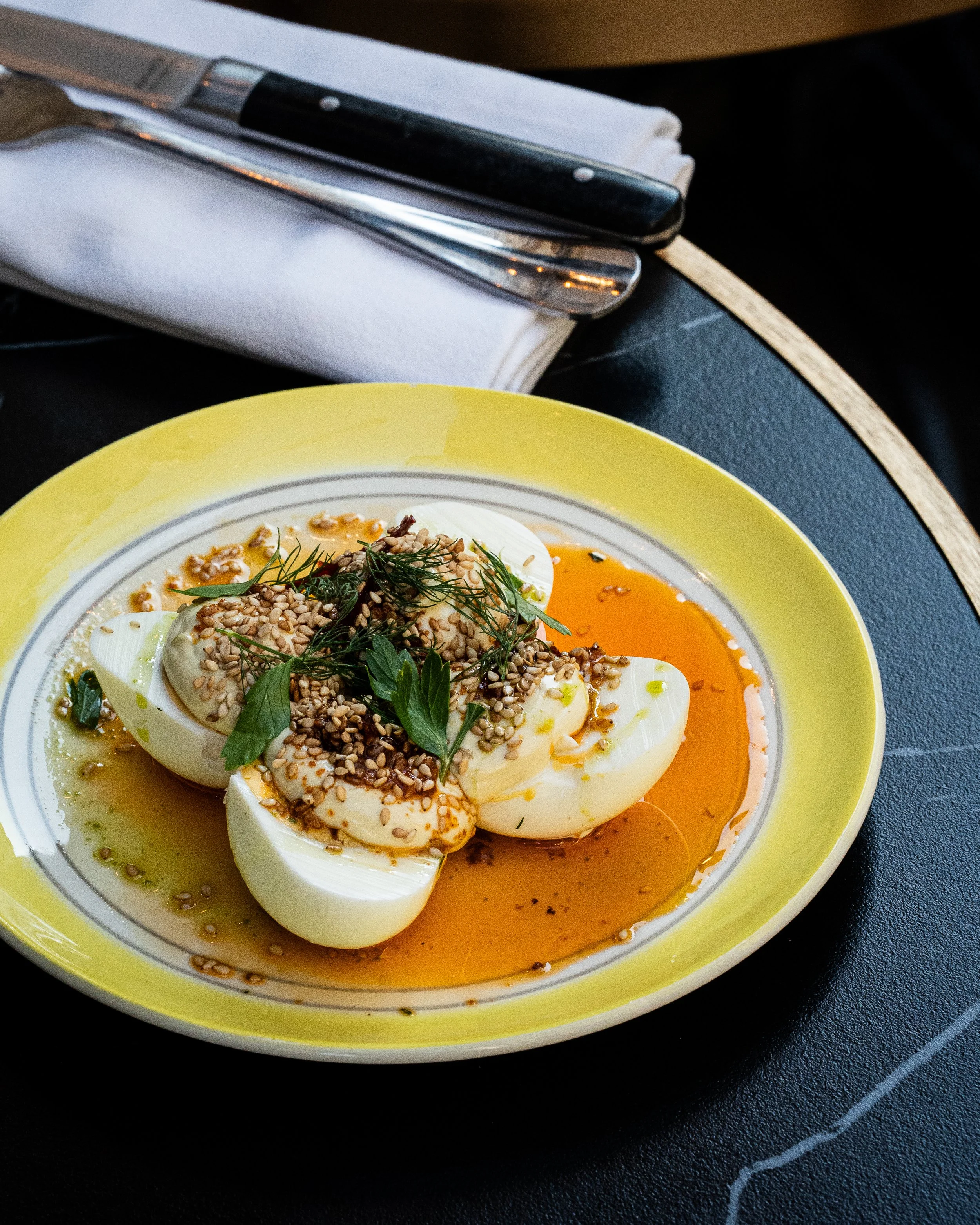 Oeuf mollet dans une assiette blanche avec des graines de sésame, des fines herbes, dans une assiette jaune, sur une table noire.