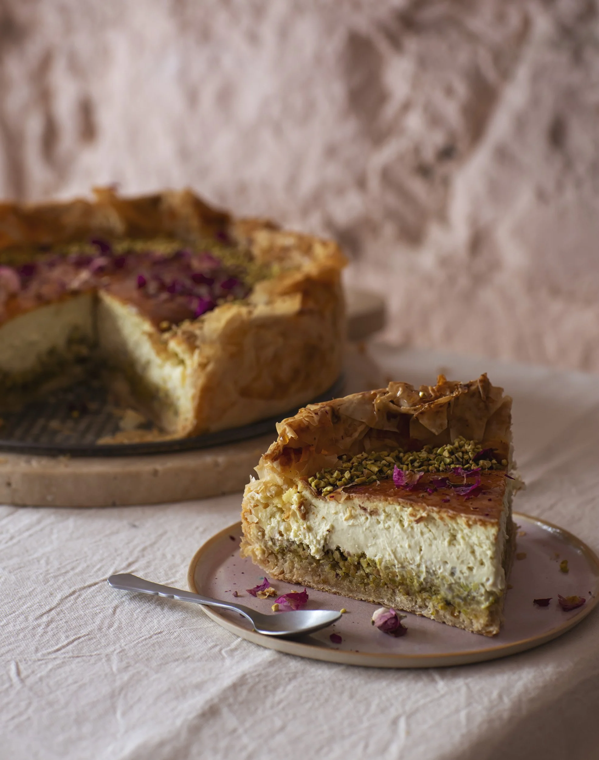 Une part de gâteau au fromage avec des pistaches et des fleurs de rose sur une assiette, une pièce du gâteau en arrière-plan.