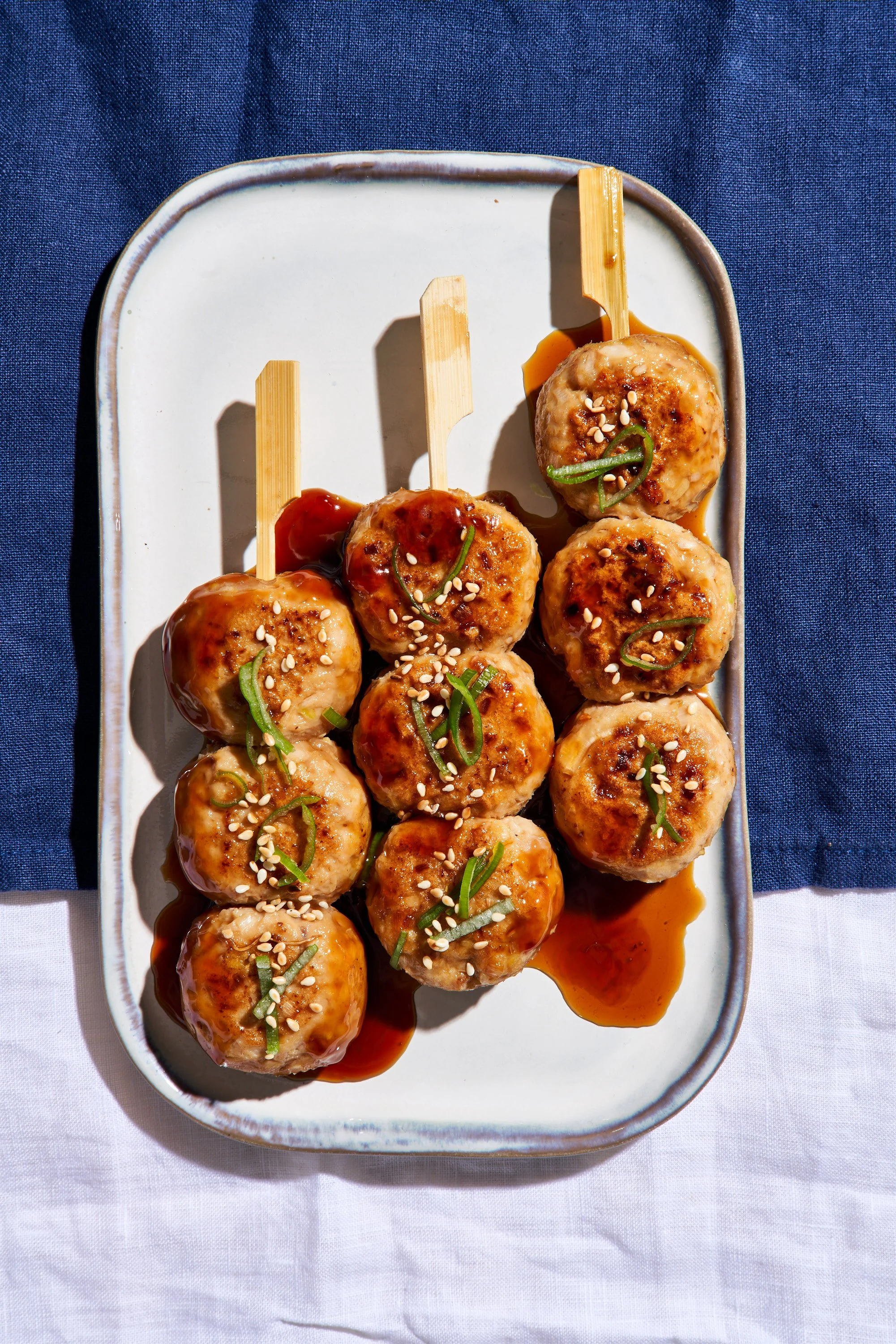 Boulette de viande avec sauce soja, garnie de ciboulette et de graines de sésame, servie sur une assiette blanche avec un fond en tissu bleu.