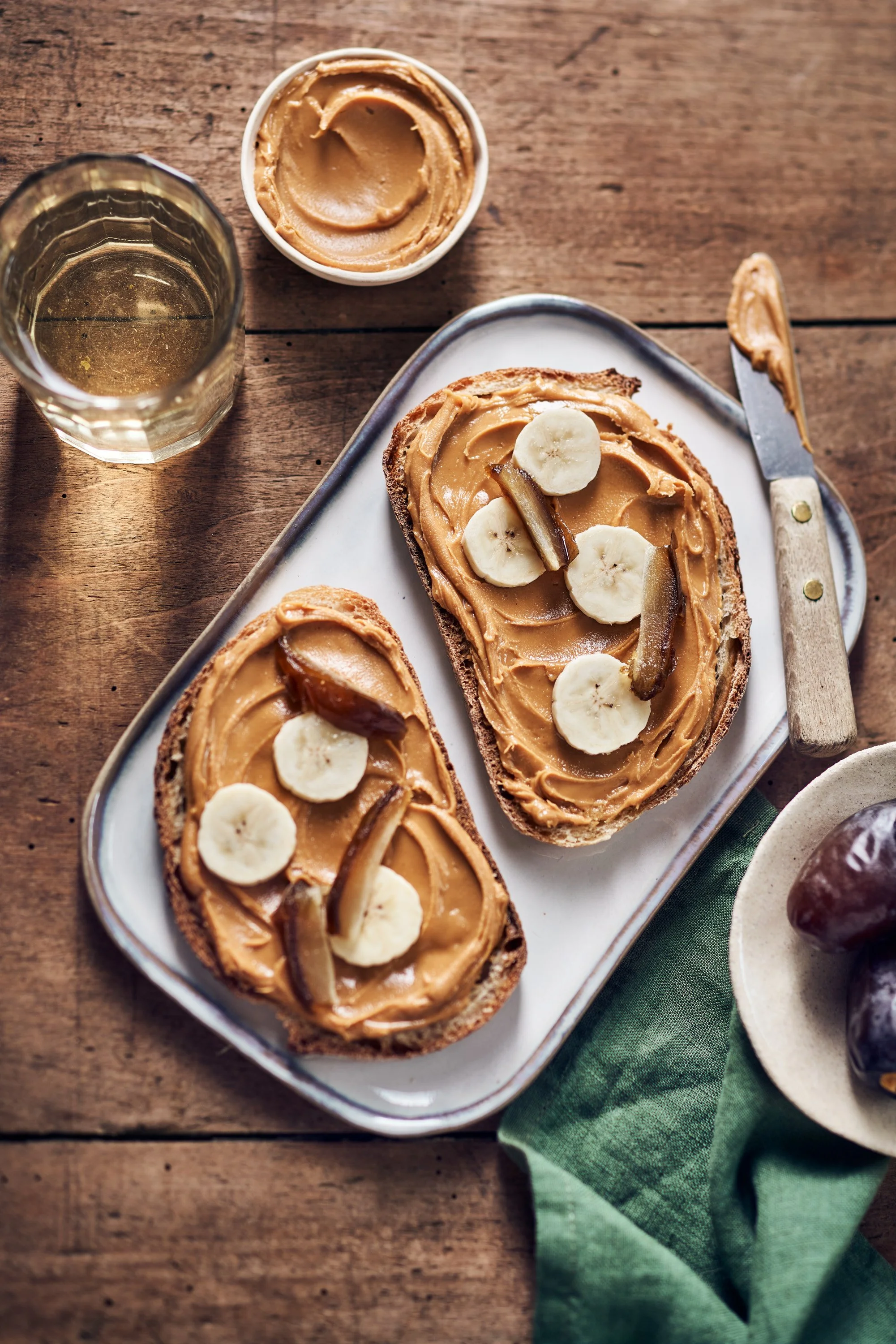 Tranches de pain avec de la beurré de beurre de cacahuète, décorées de bananes, avec une boisson et une confiture sur la table en bois, accompagnées d'un torchon vert.