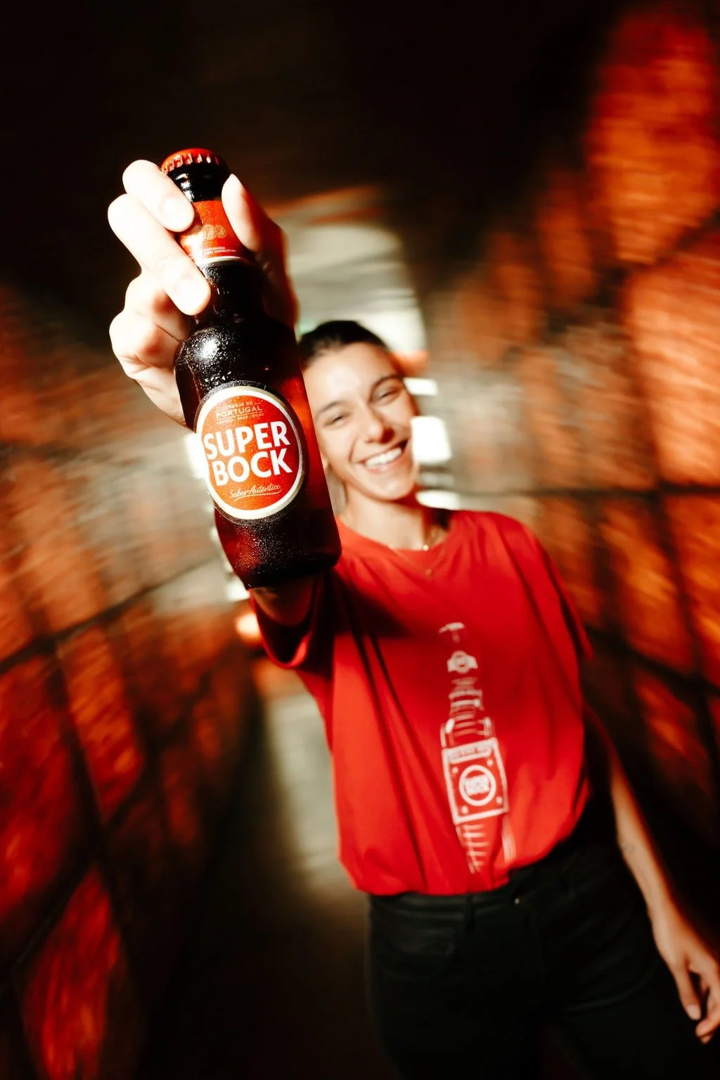 Une femme souriante tient une petite bouteille de bière 'Super Bock' dans un tunnel en briques décoratif, portant un t-shirt rouge.