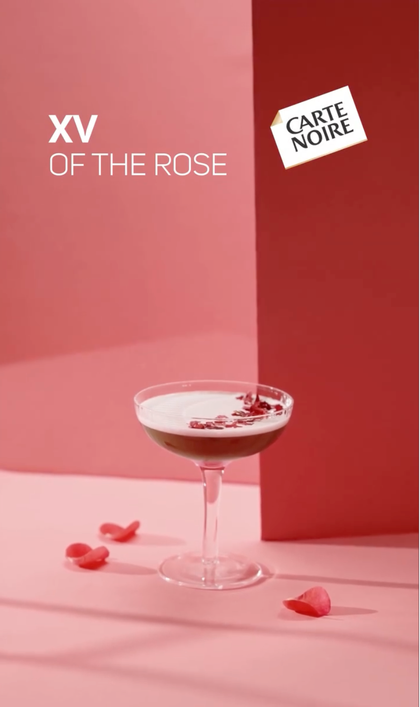 Un verre à cocktail avec une garniture rouge, posé sur une surface rose avec des pétales de rose autour, fond rose et rouge, avec du texte 'XV of the Rose' et une étiquette 'Carte Noire'.