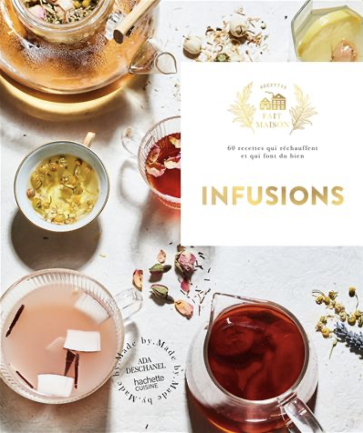 Un livre intitulé 'Infusions' entouré de plusieurs tasses de différentes couleurs de tisanes ou d'infusions, accompagnées de fleurs séchées et de fleurs fraîches, avec une pièce de monnaie et un mot inscrit 'Made by Ada Deschanel'.