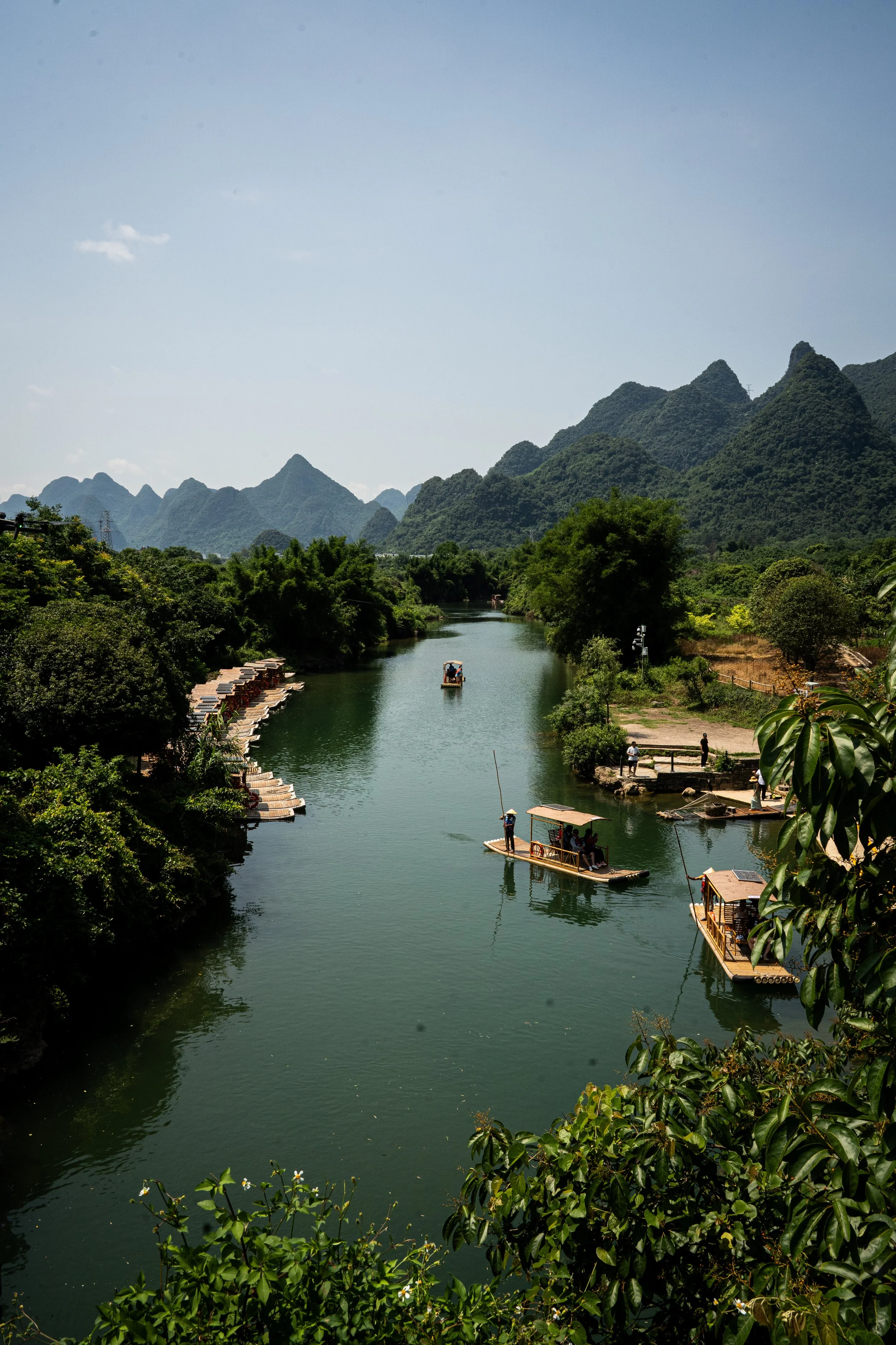 Scène d'une rivière traversant une région montagneuse, avec plusieurs bateaux en bois transportant des passagers et des touristes, entourée de végétation dense et de montagnes verdoyantes sous un ciel clair.