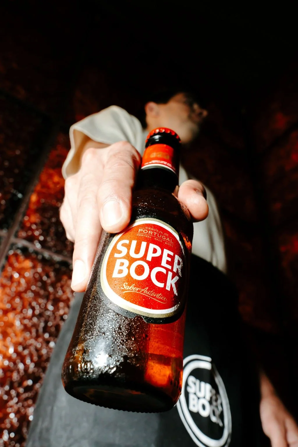 Une personne tient une bouteille de bière Super Bock, avec un fond sombre et une surface mouillée ou couverte de gouttelettes d'eau.