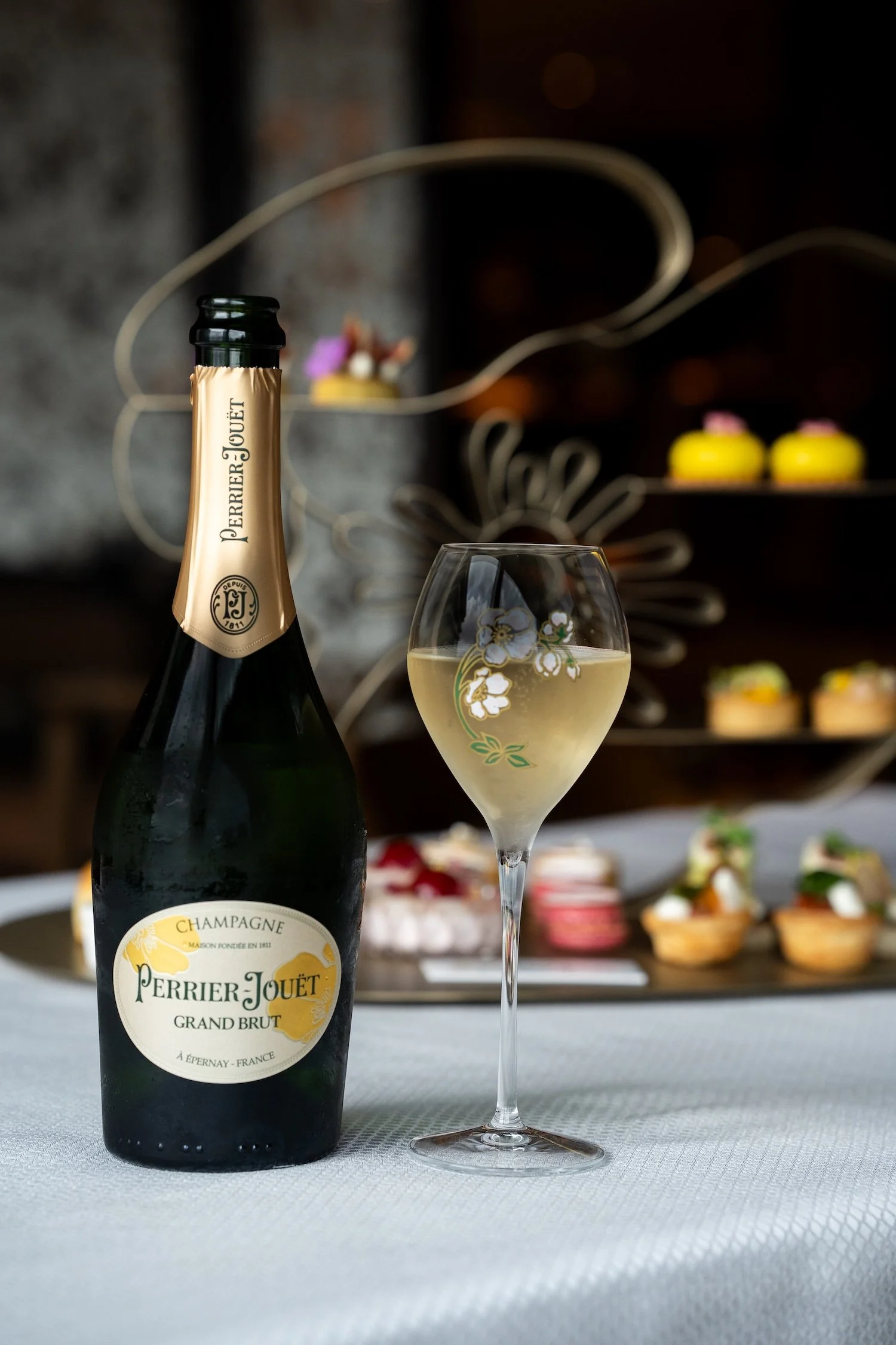 Une bouteille de champagne Perrier-Jouët Grand Brut avec un verre de champagne à côté, posé sur une table, avec un décor de pâtisseries en arrière-plan.