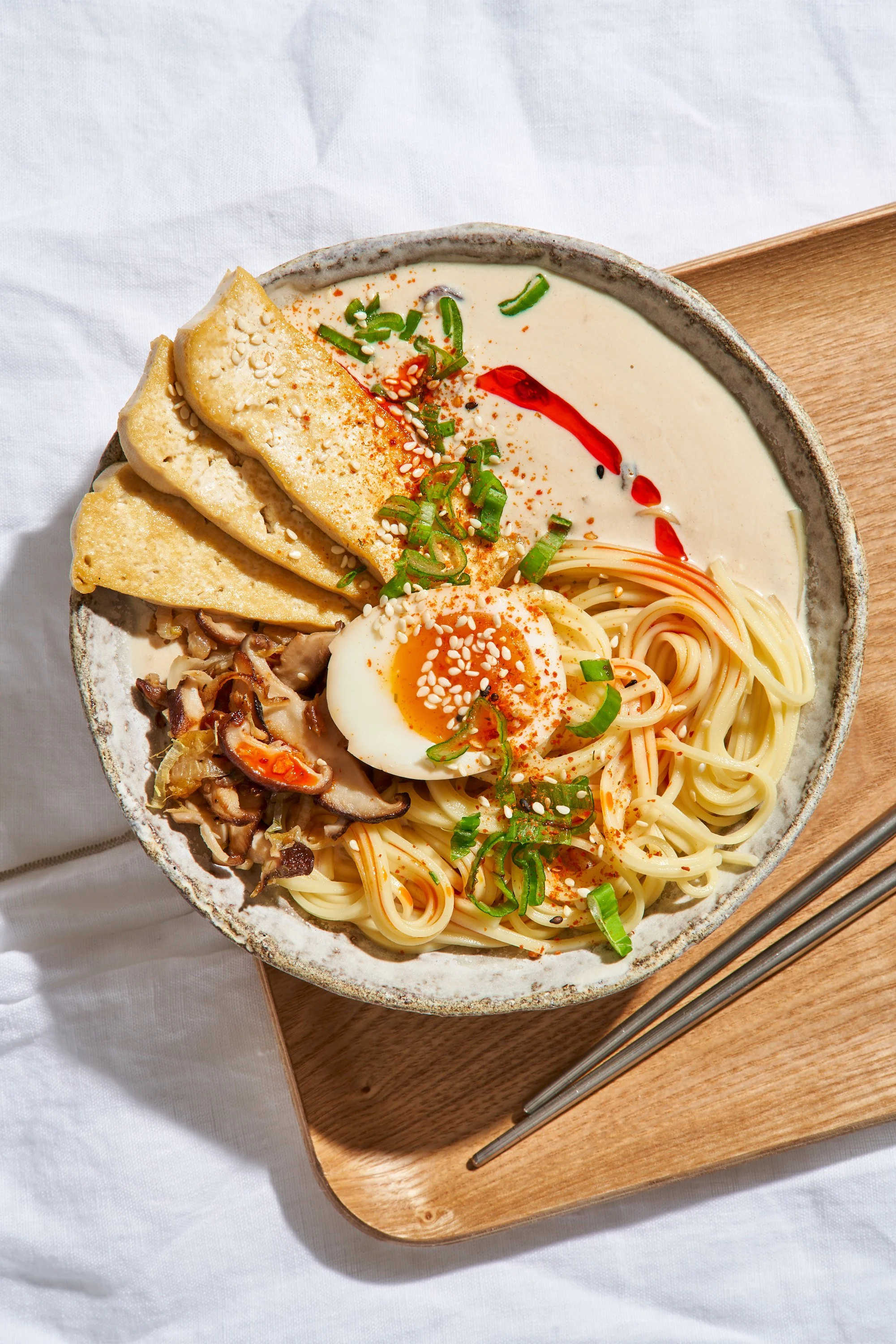 Un bol de ramen avec œuf mollet, nouilles, champignons, tofu, oignons verts, et sauce rouge, posé sur un plateau en bois.