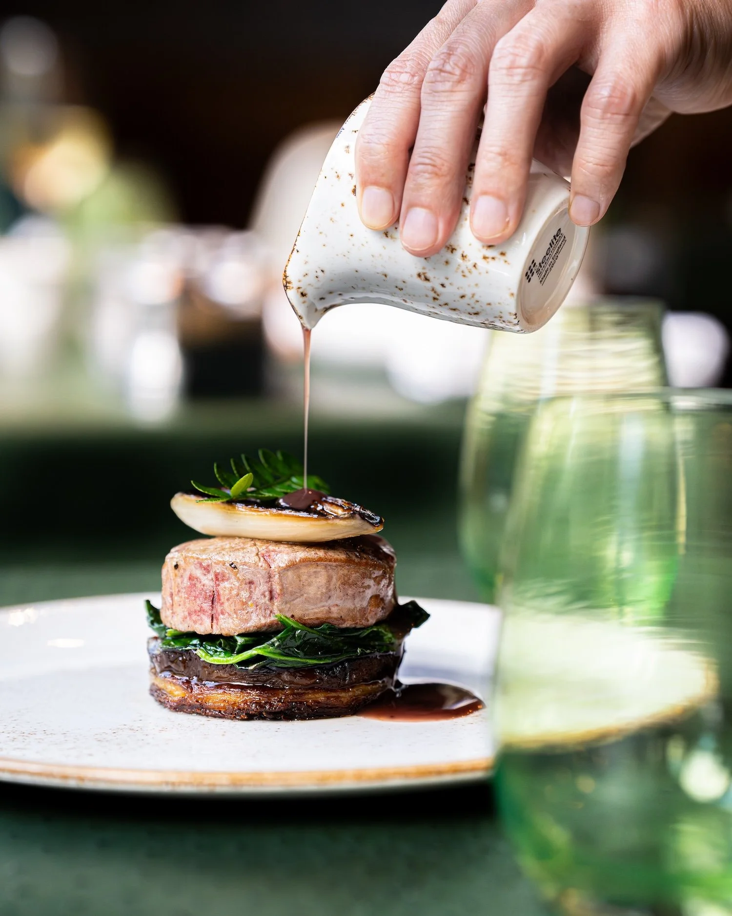 Une main verse une sauce dans un plat de viande et légumes, avec un arrière-plan flou de verres et de table.