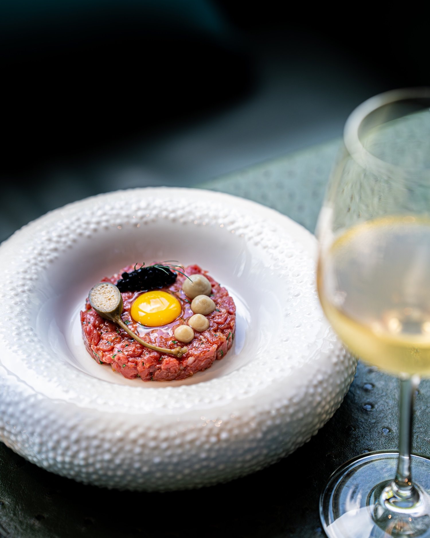 Tartare de viande avec un œuf au centre, présenté dans une assiette blanche avec un verre de vin blanc à proximité.