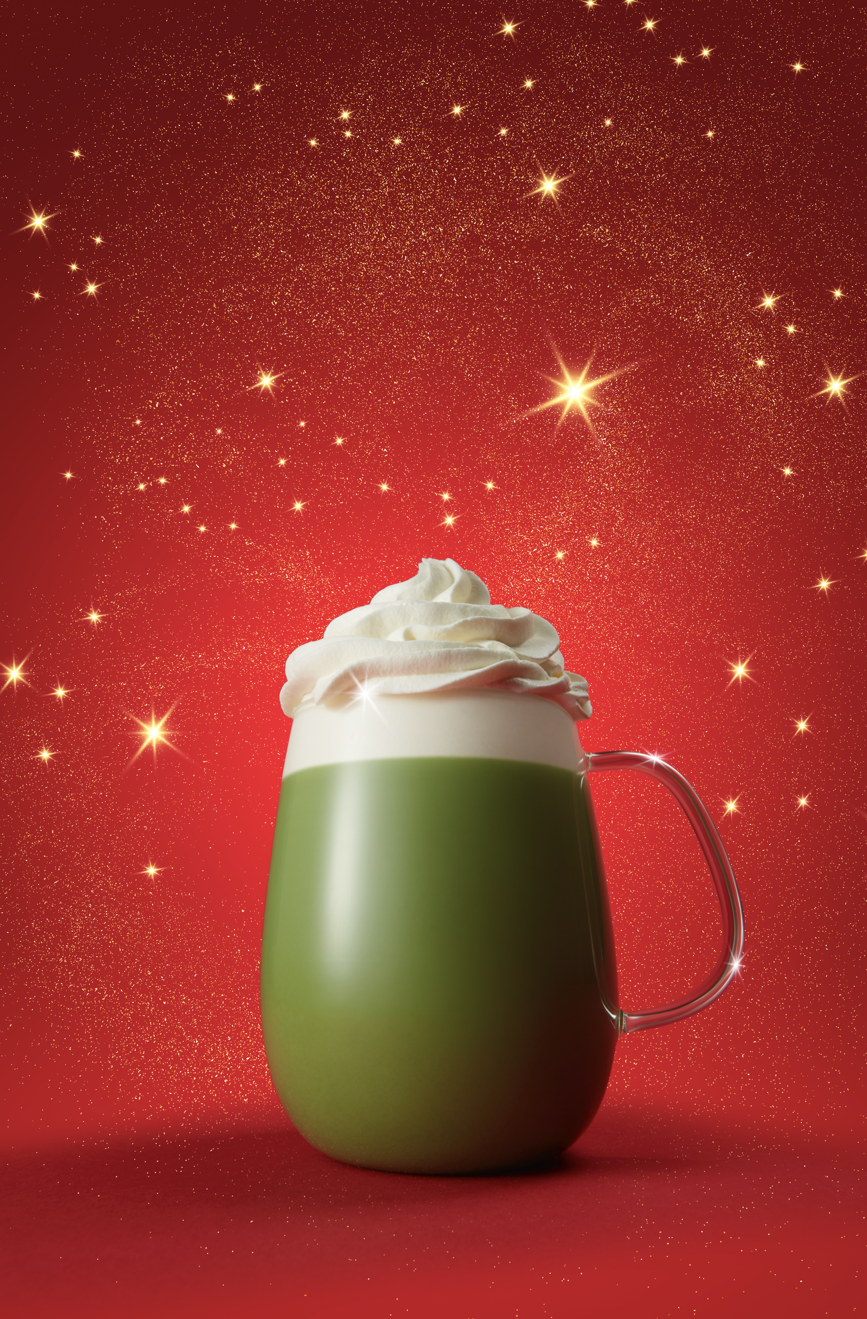 Une tasse verte avec une poignée en verre, remplie de chocolat chaud et de crème fouettée, sur fond rouge avec des étincelles dorées et des étoiles scintillantes.